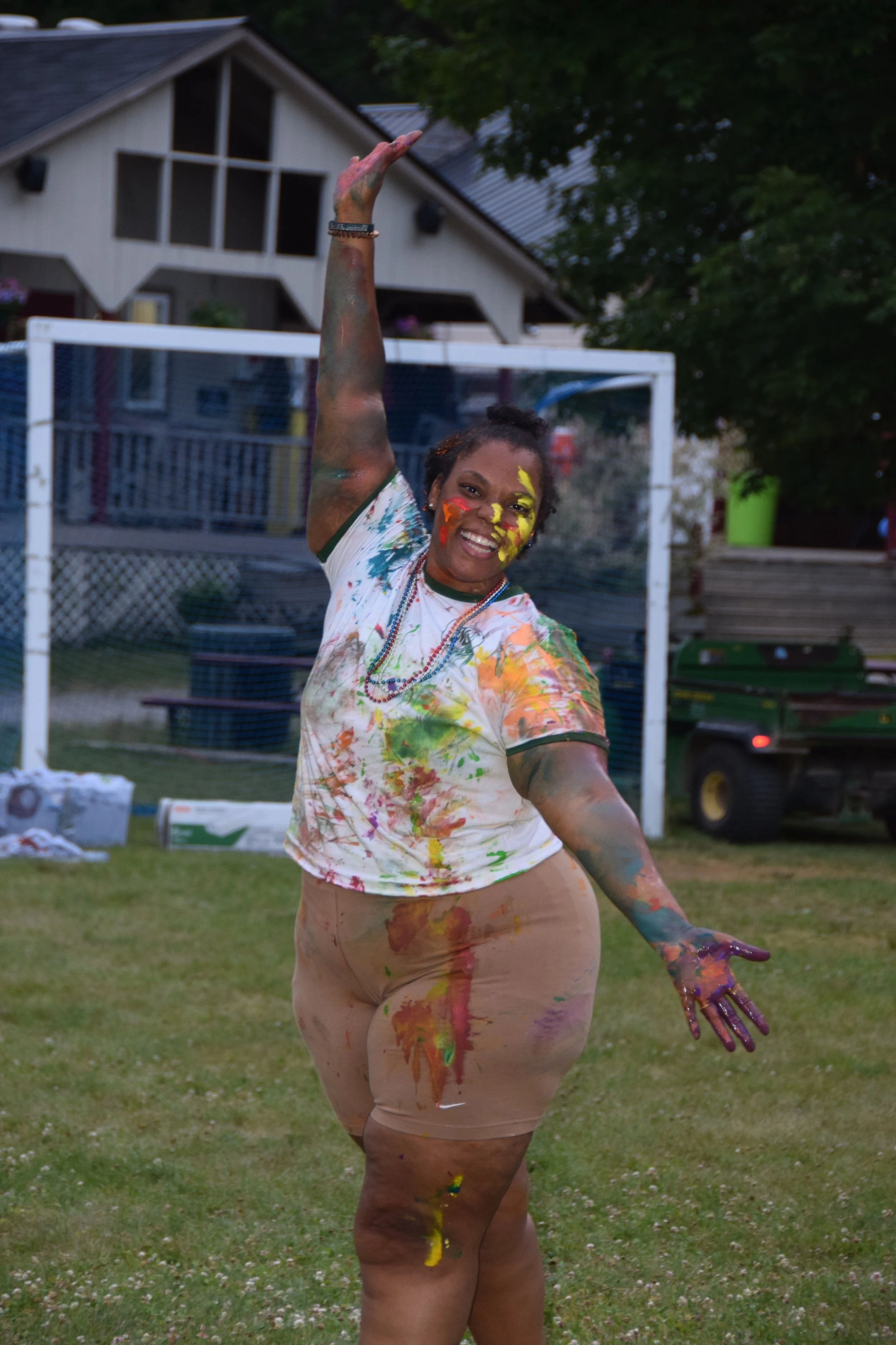 A joyful woman covered in multicolored paint, smiling and dancing outdoors on grass with a house, trees, and gardening equipment in the background.