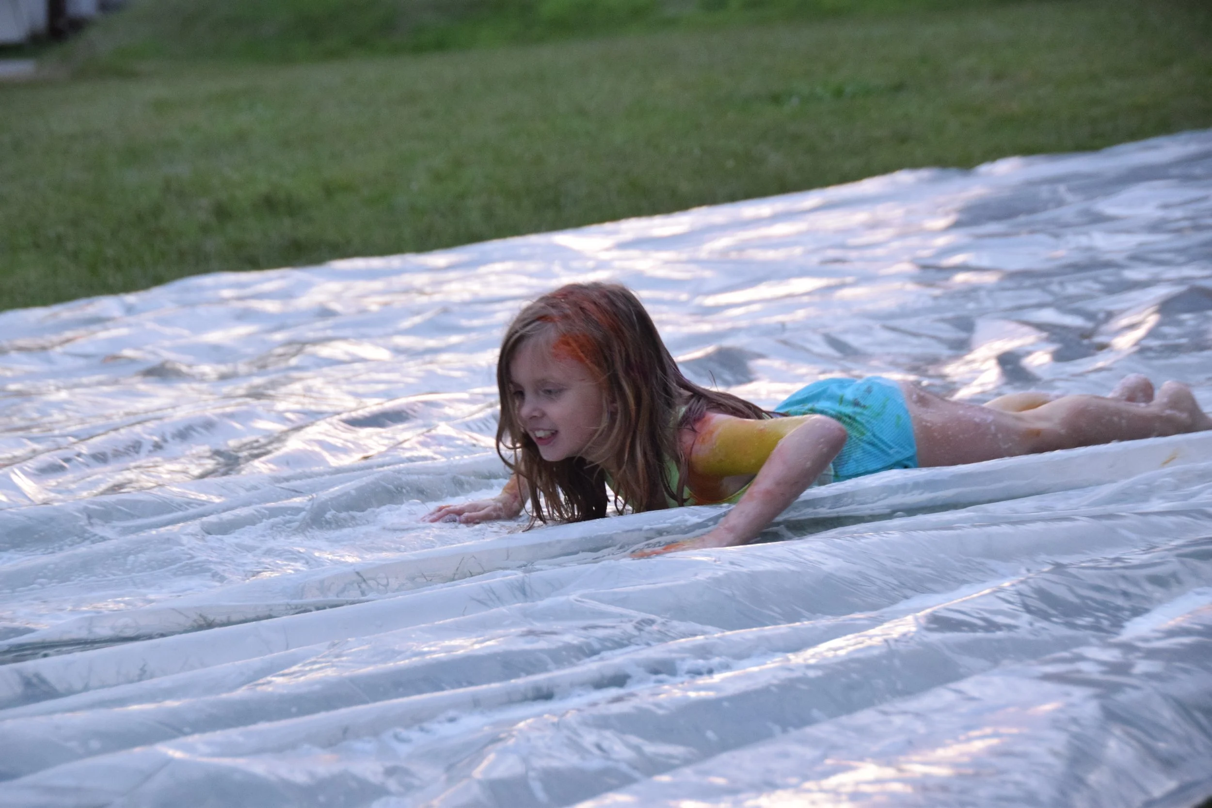 A young girl with orange and brown hair lying on a plastic slip and slide lawn surface, wearing a colorful swimsuit, and having fun.