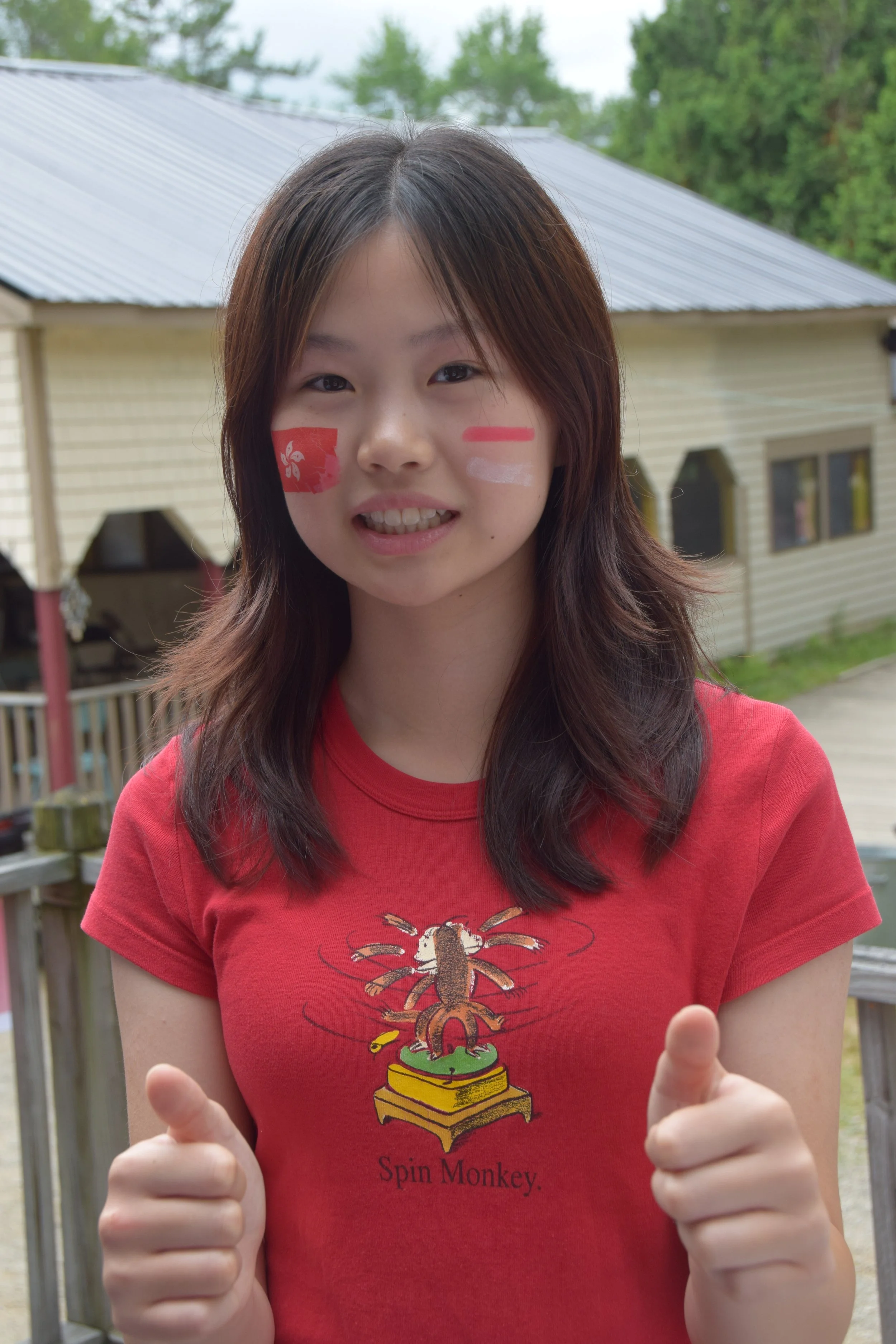 Young woman outdoors with face paint and a red T-shirt, showing two thumbs up and smiling at the camera.