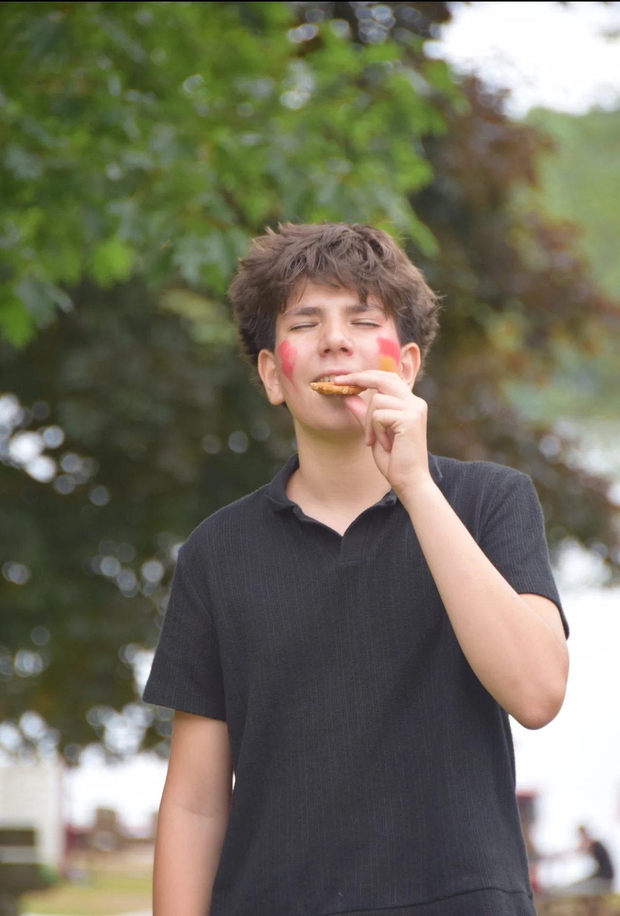 Young person with face paint, eating a cookie outdoors with trees in the background.
