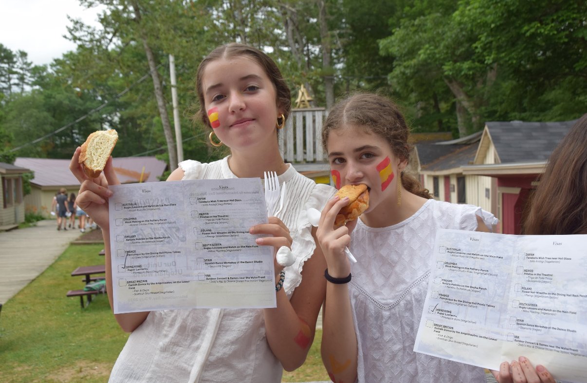 Two young girls with face paint of Spain's flag holding papers and eating snacks outdoors.