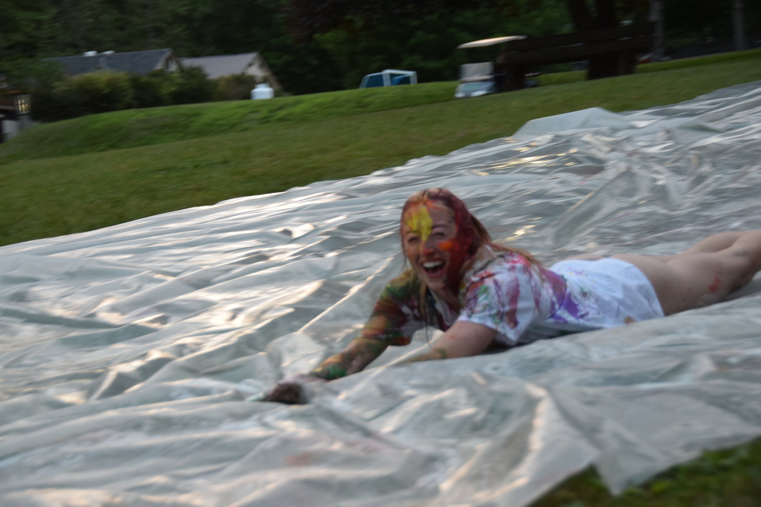Person smiling and covered in colorful paint on a slip and slide in a backyard.
