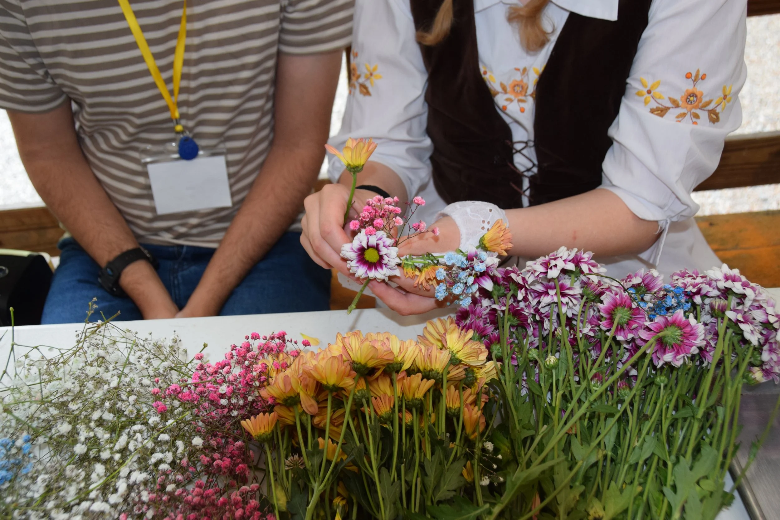 A person arranging a variety of colorful flowers on a table while another person looks on. The flowers include pink, orange, white, blue, and purple blooms in a floral arrangement.