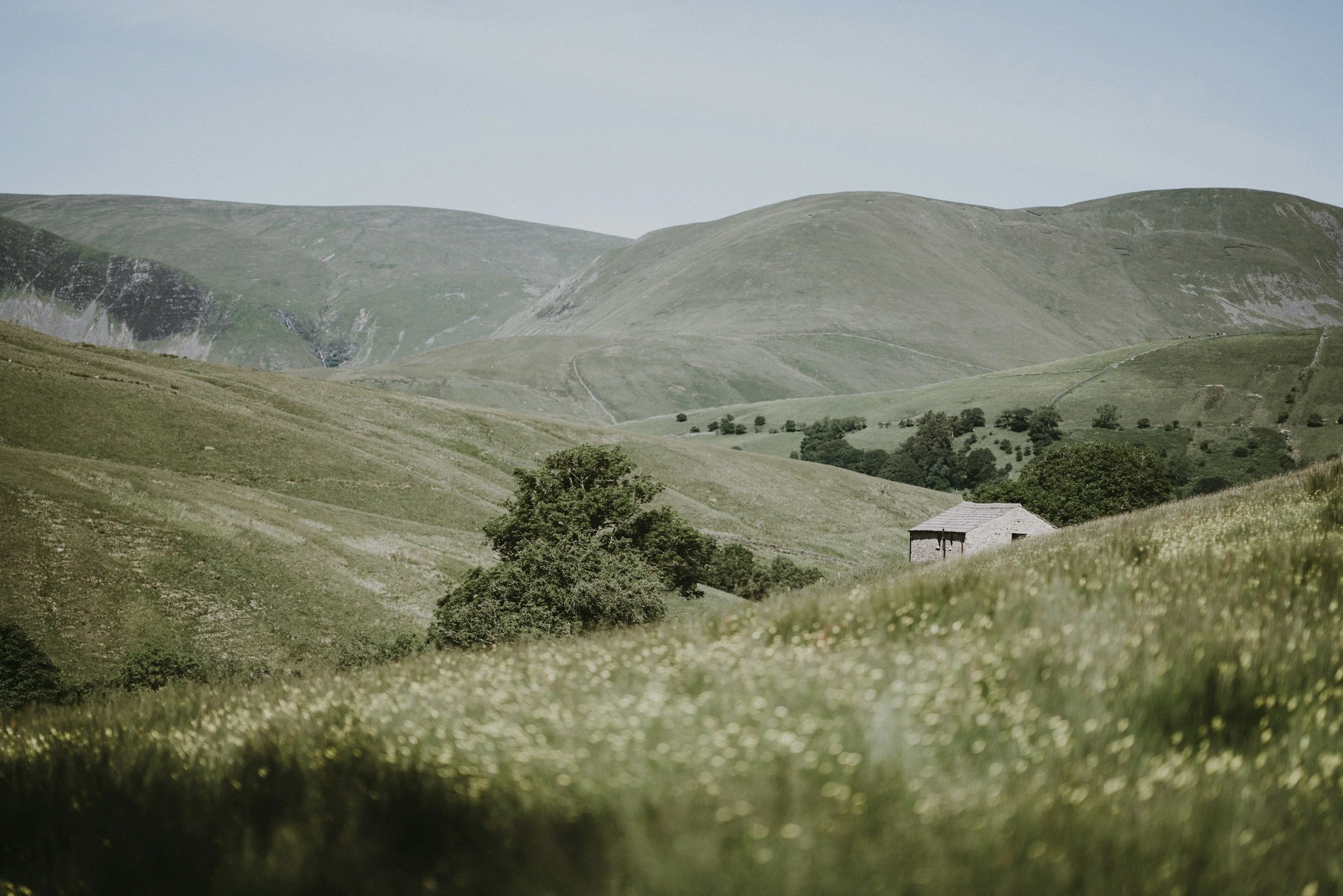 A landscape of rolling green hills with a small stone building in the foreground and a partly cloudy sky.