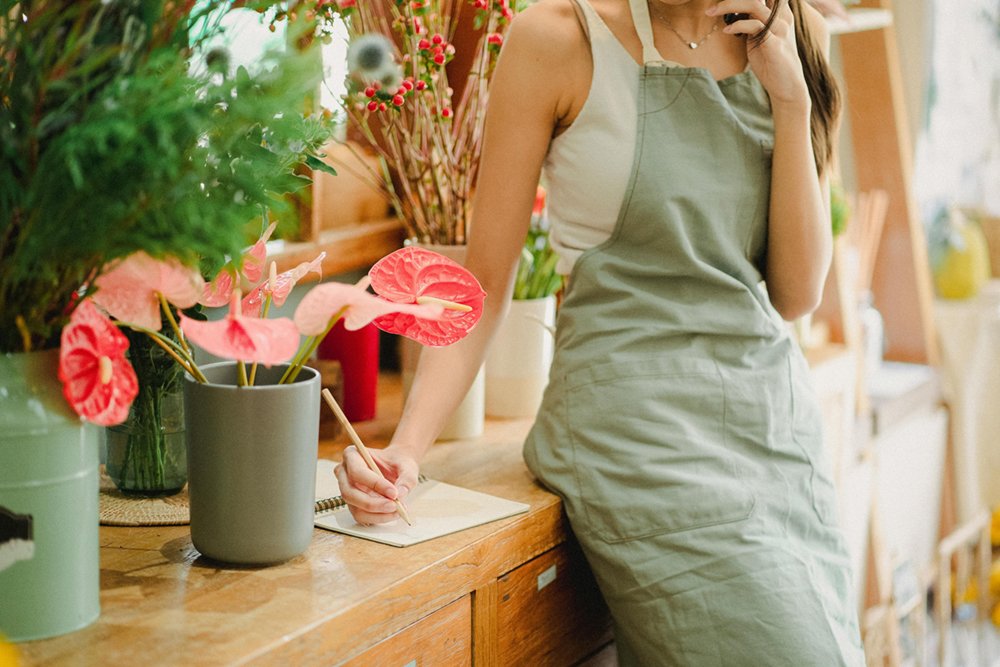 A woman florist on the phone, taking notes, while leaning against a counter with flowers around her. She his a business woman.