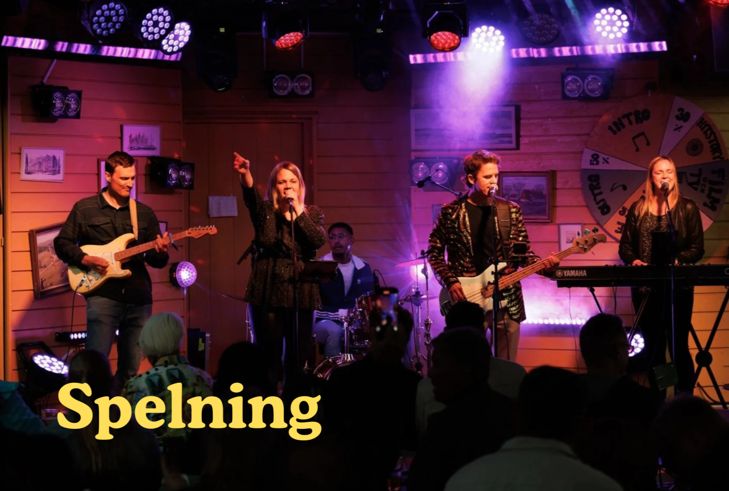 En musikband som spelar live på en scen med trädväggar och pulserande ljus. Fyra musiker: en man med elgitarr, en kvinna med mikrofon, en man med elbas, och en kvinna med keyboard. En publik sitter och tittar på.