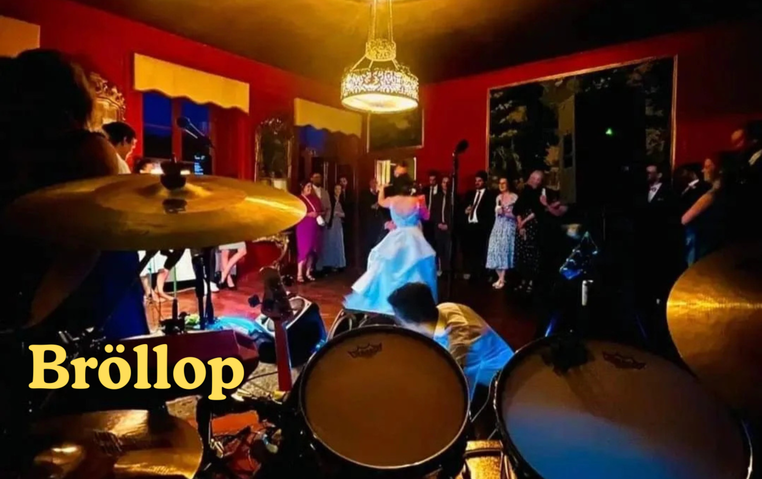 Kvällssdukning med ett bröllopsparty på en inomhusclub. Bruden i blå klänning dansar, omringad av gäster i formella kläder. Tävlar på en scen med musik och en trummis synlig i förgrunden.