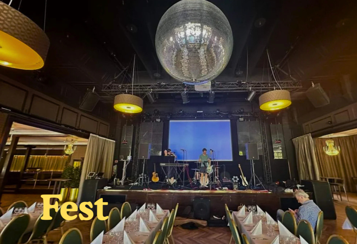 En festlokal med ett scenställe där två musiker står framför en stor skärm. Det finns ett disco kul på taket och flera bord är dukade med glas, bestick och servetter. Personen i förgrunden tittar på sin mobiltelefon.
