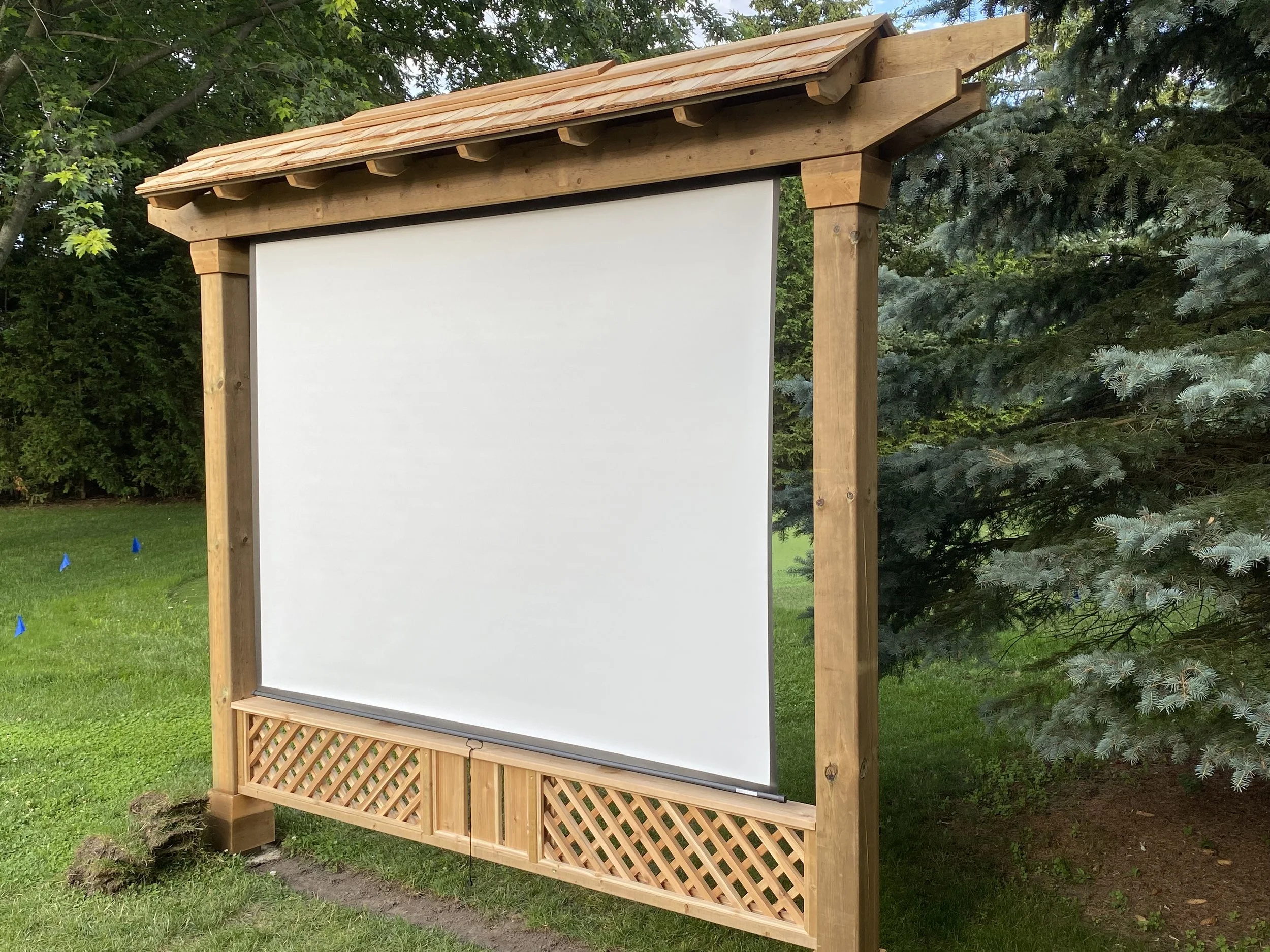 An outdoor projection screen with a wooden frame and lattice at the bottom, set up on a grassy area surrounded by trees.