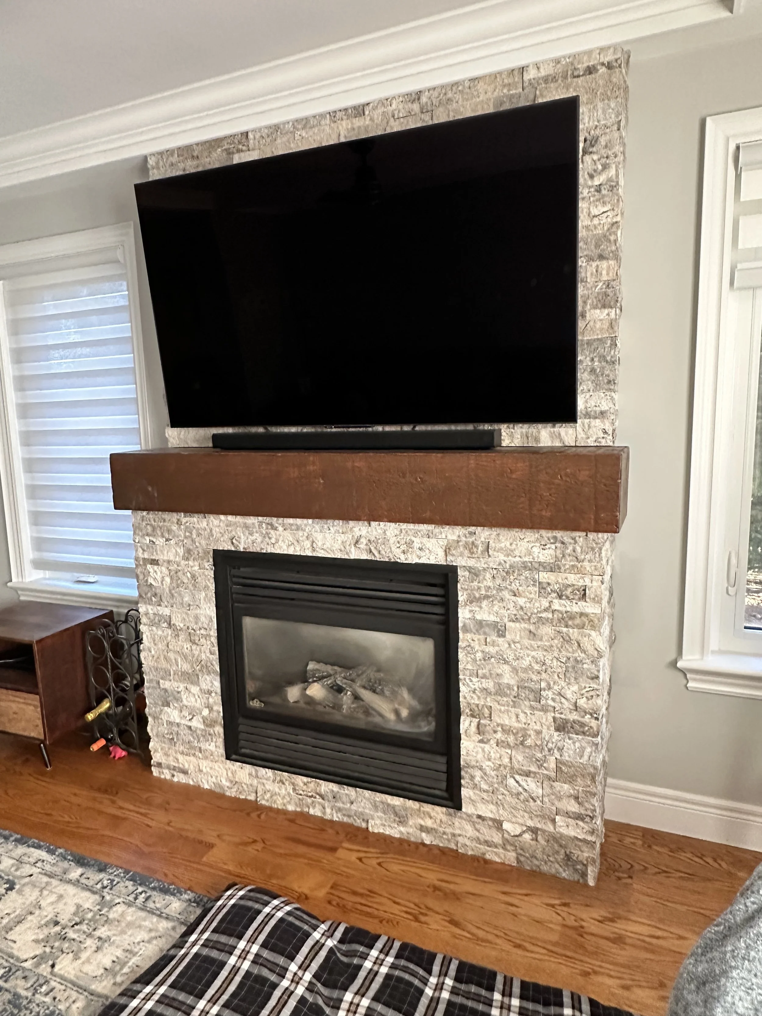 A living room fireplace with a mounted flat-screen TV above it, surrounded by gray stone, with a wooden mantel. To the left, there is a window with horizontal blinds, and a small wooden wine rack is beside the fireplace. Part of a patterned rug and f