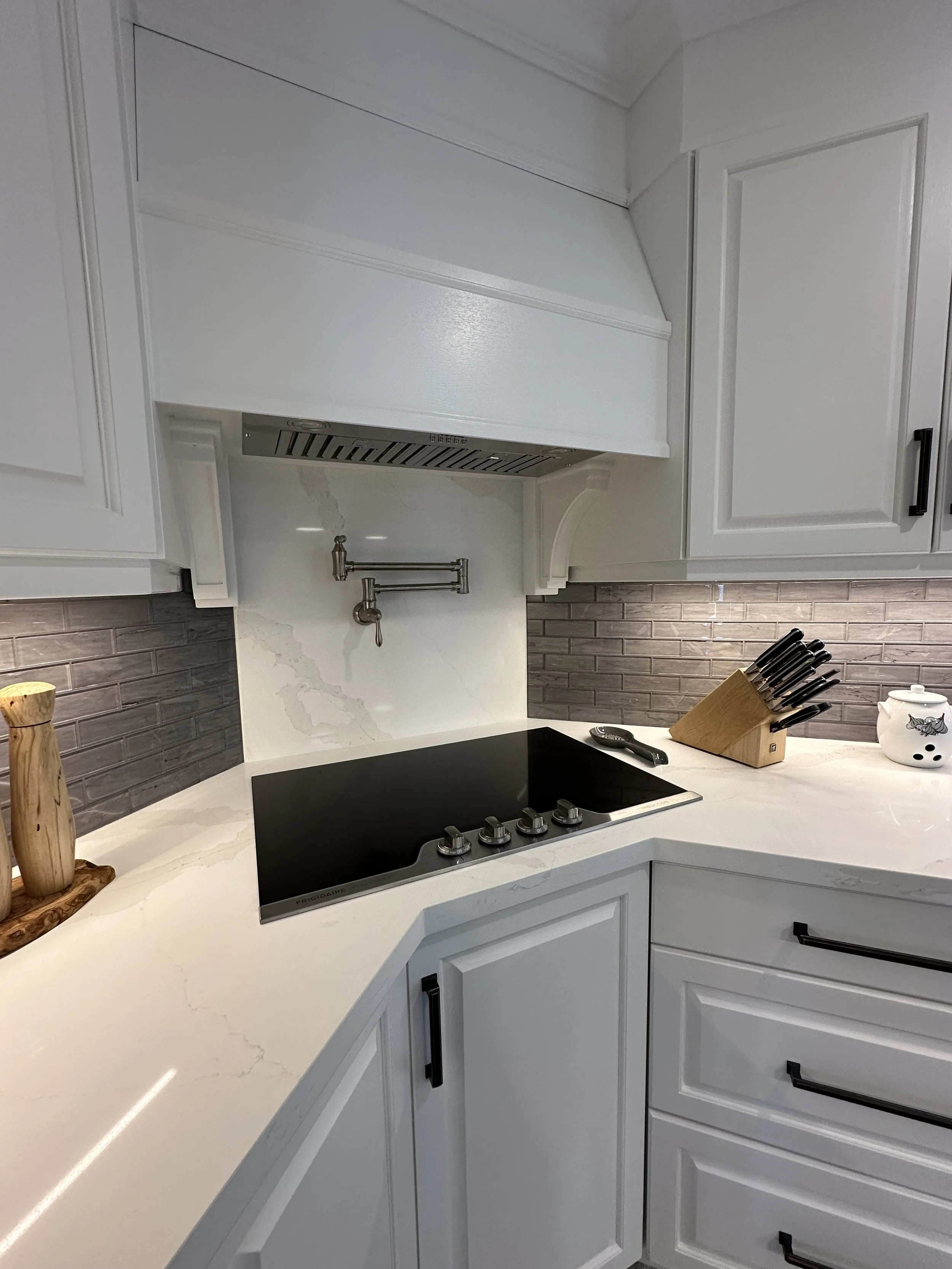 A modern kitchen with white cabinets, a black electric cooktop, a knife block on the countertop, and a gray brick backsplash.