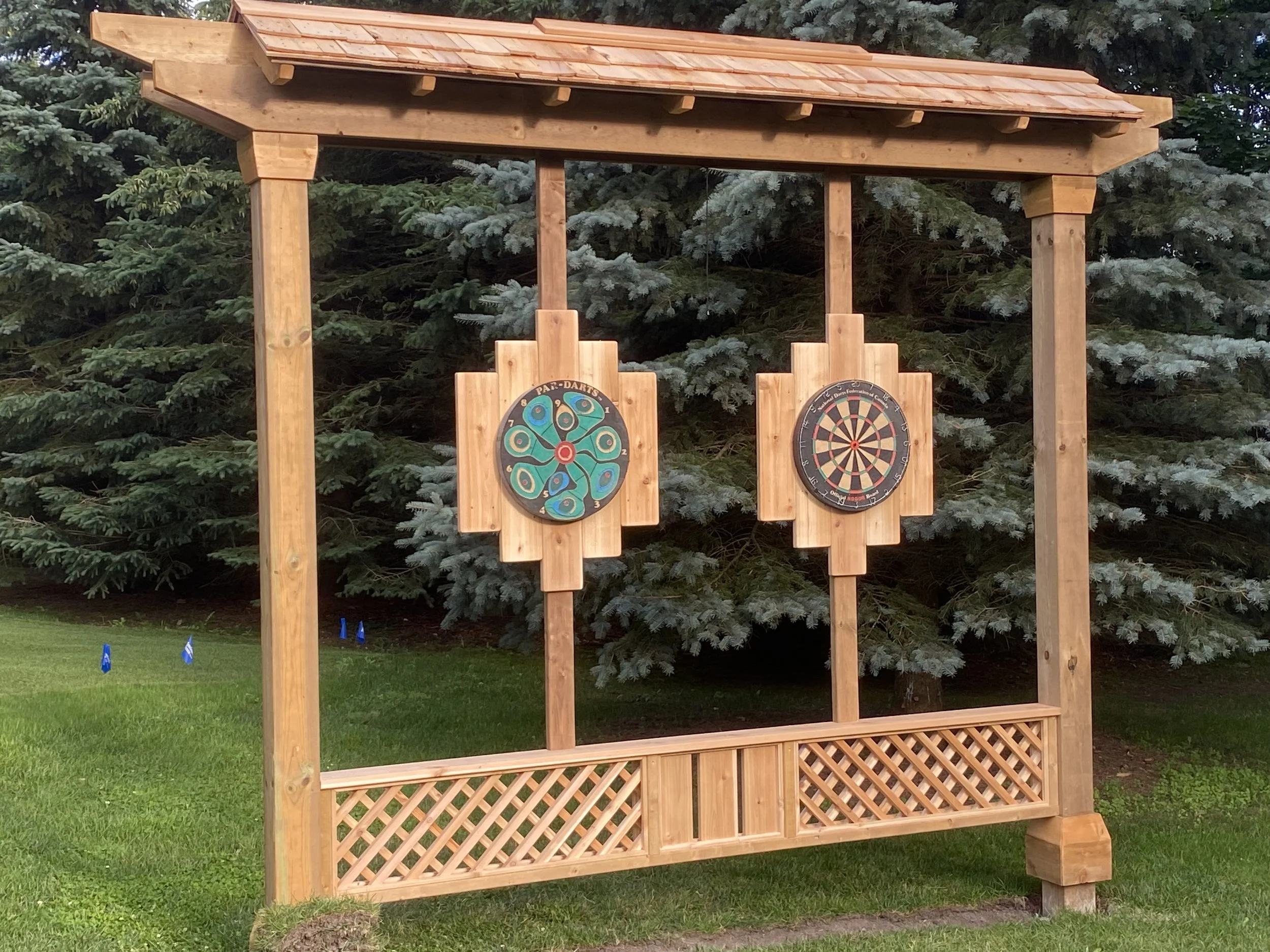 A wooden outdoor game station with a dartboard on the right and a colorful circular game on the left, set against a backdrop of evergreen trees.