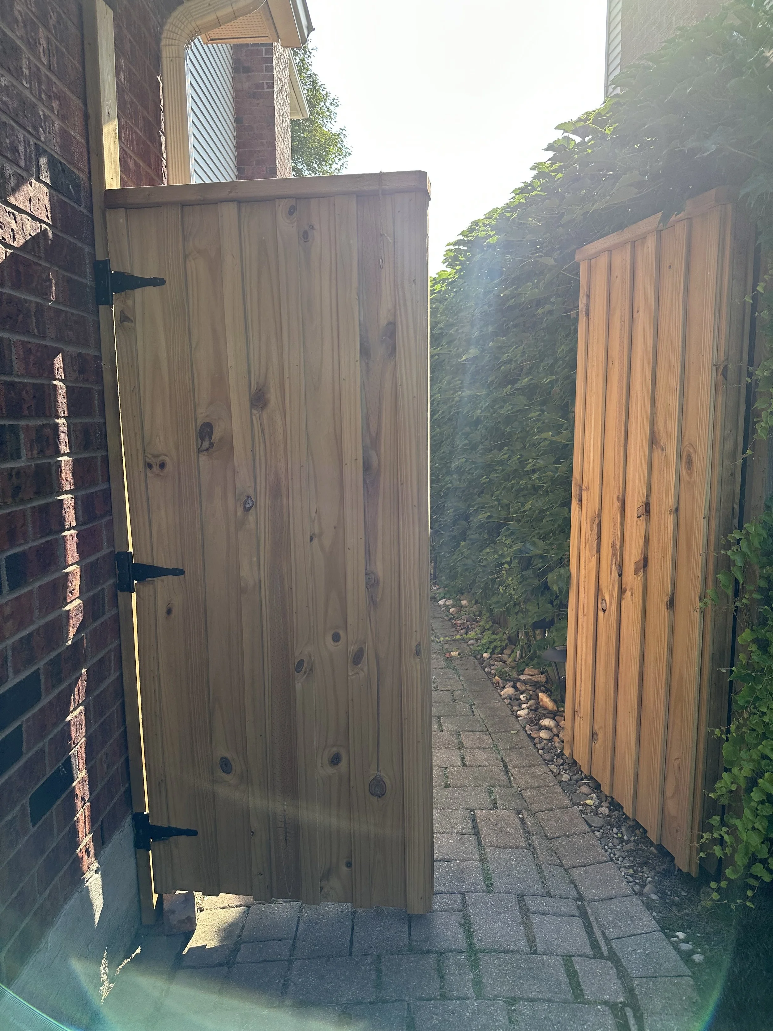 View of a small backyard passageway with a custom wooden gate open to outdoor space. The path is paved with brick stones, and there is greenery on the right side. Part of a brick house and a neighboring house are visible in the background, with sunli