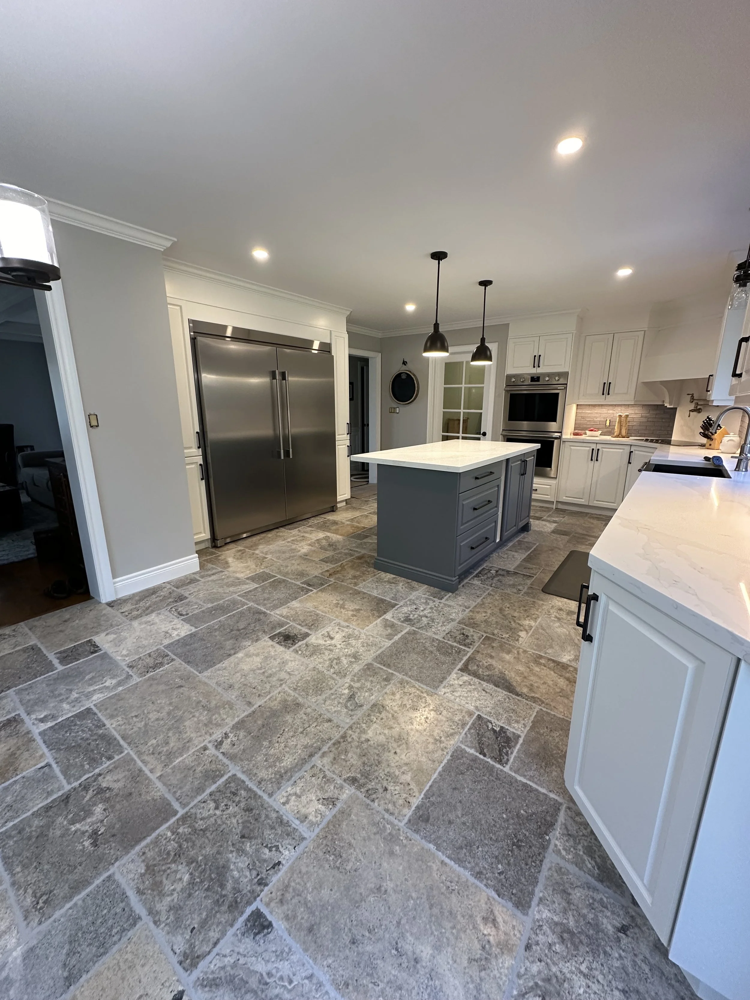 Modern kitchen with white cabinets, gray island, stainless steel refrigerator, double ovens, and tiled floor.