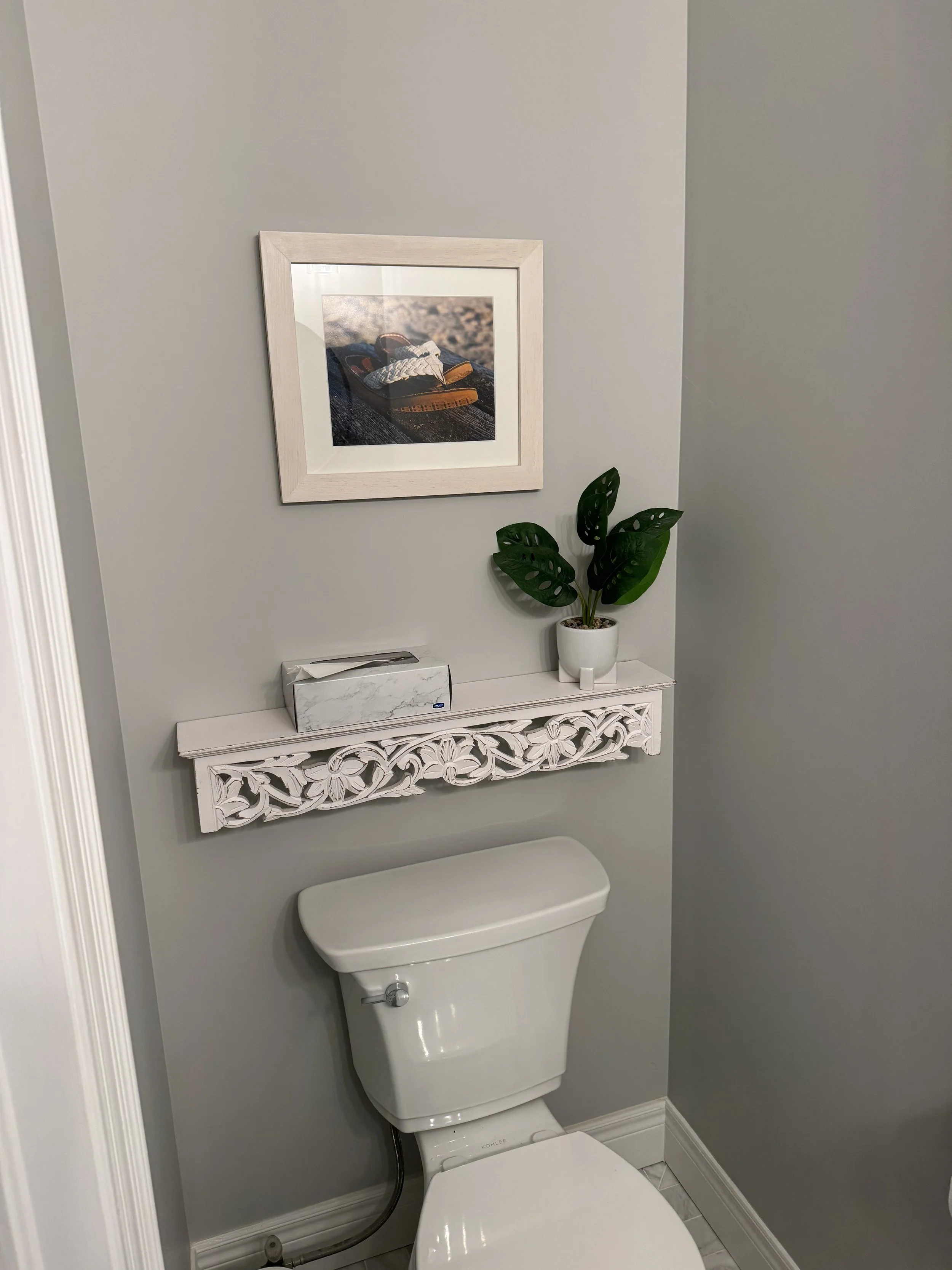 A bathroom with a white toilet, a decorative white shelf, a potted plant, and a framed photograph of sandals on a beach.