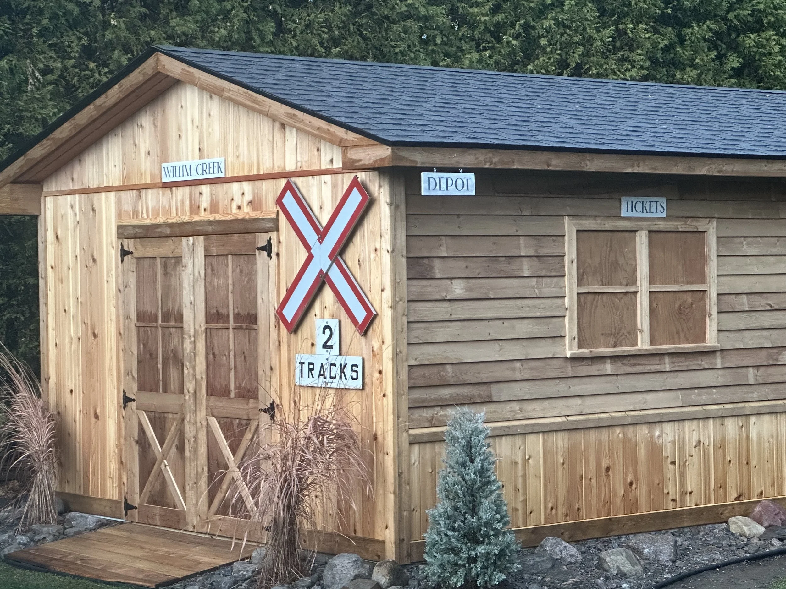 A wooden building with signs labeled 'WILLIAM CREEK', 'DEPOT', and 'TICKETS'. It has a large railroad crossing sign with '2 TRACKS' on it, and a double door. There is a small shrub and rocks in front of the building.