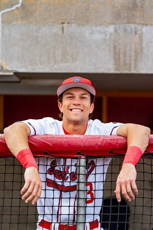 A baseball player in a white and red uniform and cap, smiling and leaning on a dugout railing with arms resting on top.