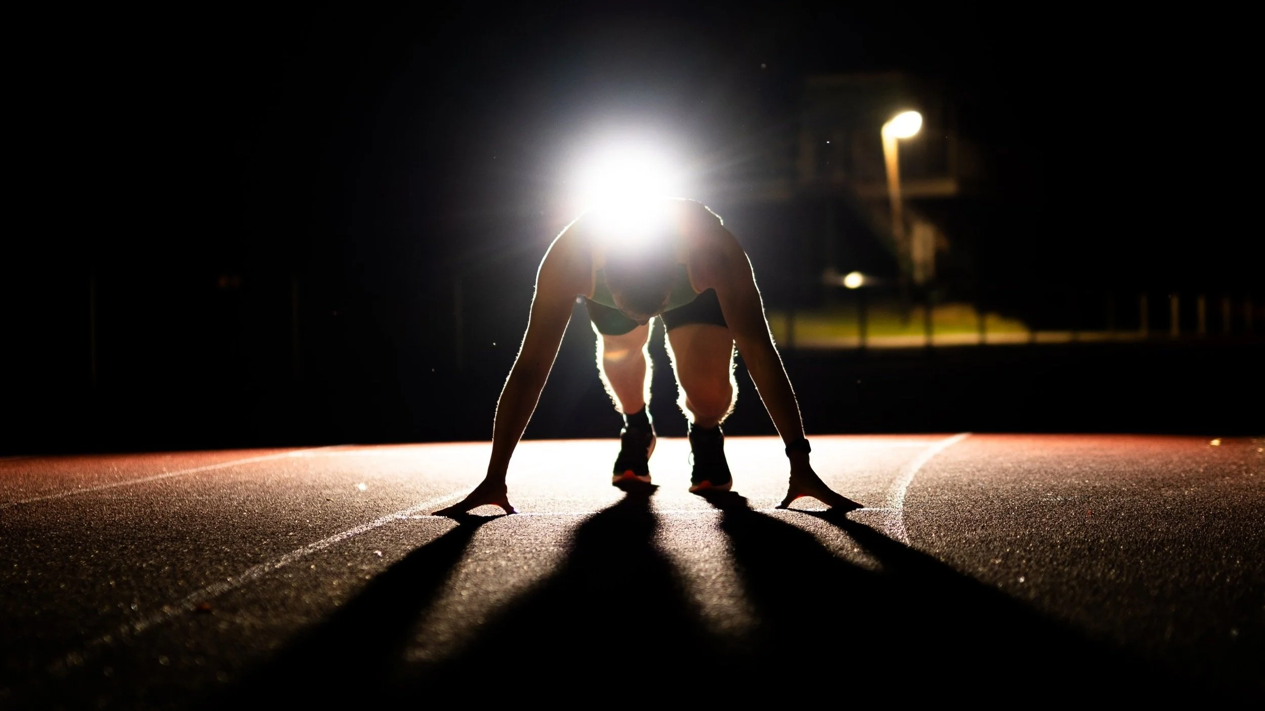 Eine Person in Startposition auf einer Laufbahn bei Nacht, beleuchtet von einem hellen Licht im Hintergrund.