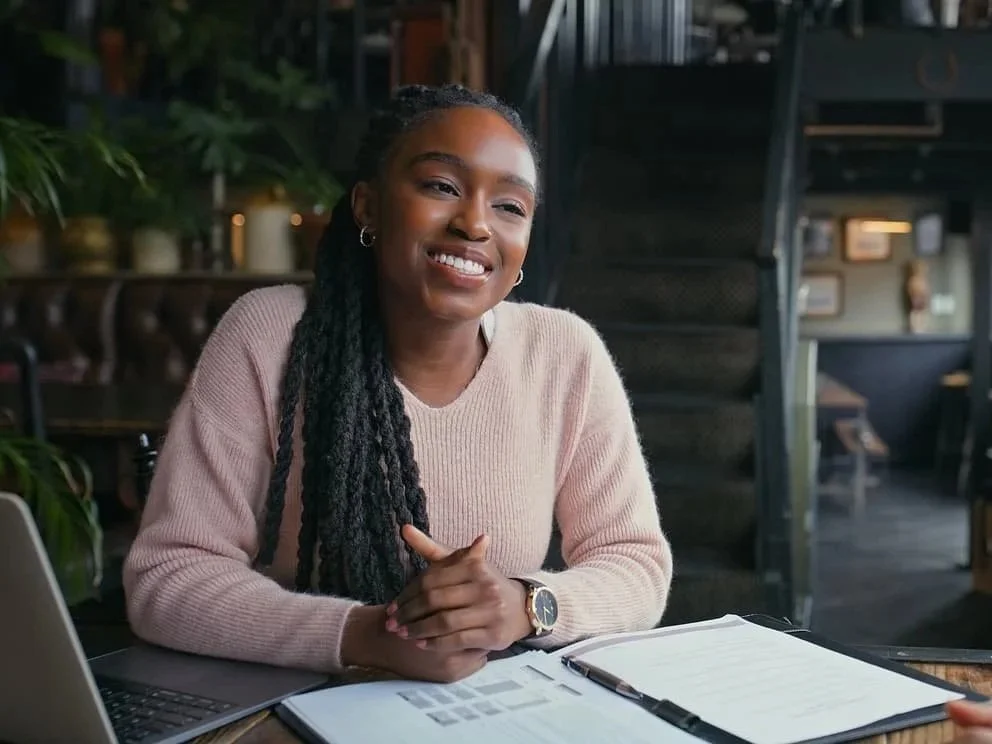 A woman with long braids smiling while sitting at a table with a laptop and notebooks in a cozy cafe setting.