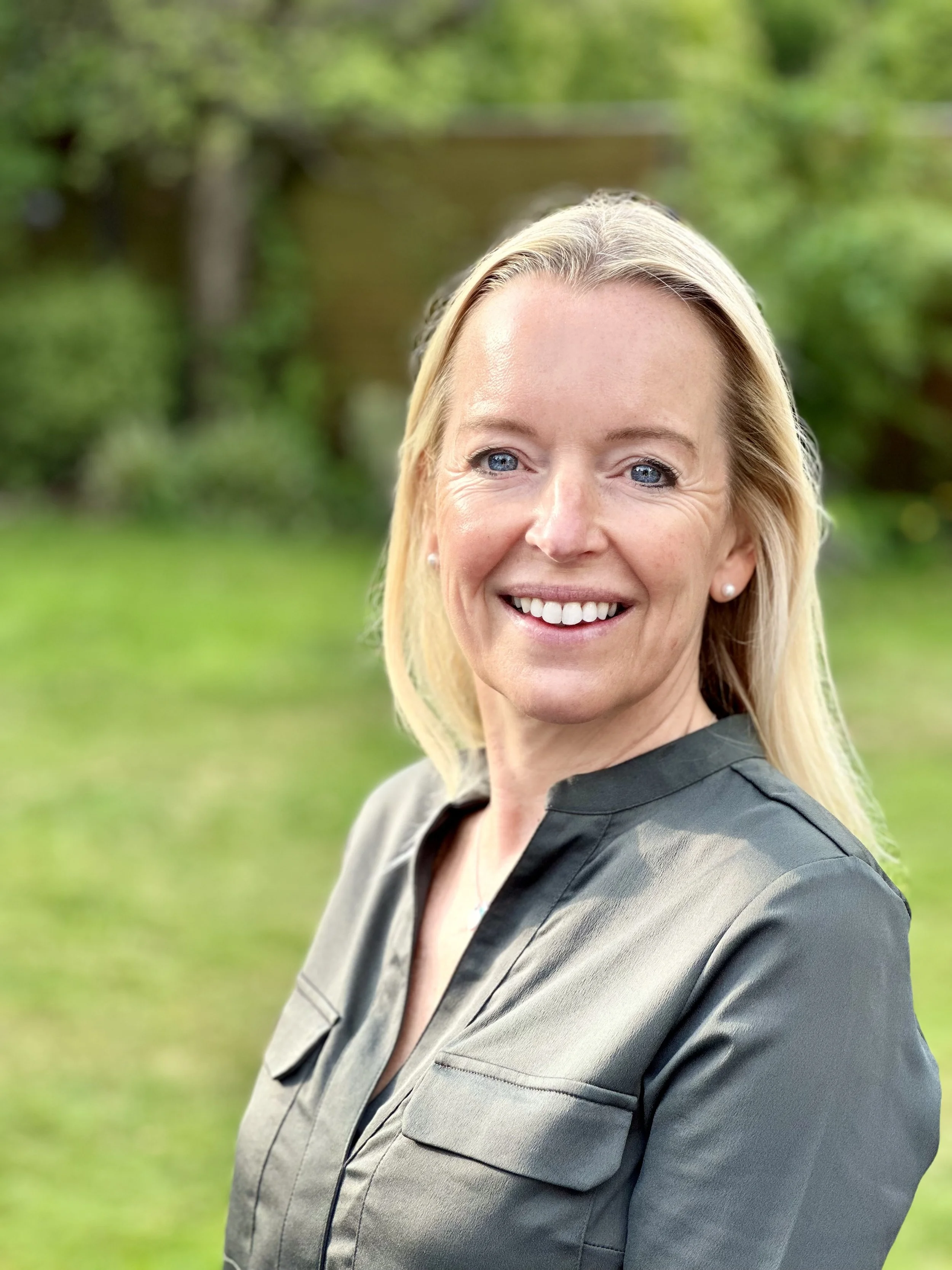 Close-up of a smiling woman with blonde hair, blue eyes, wearing a dark gray blazer, standing outdoors with a blurred green park background.