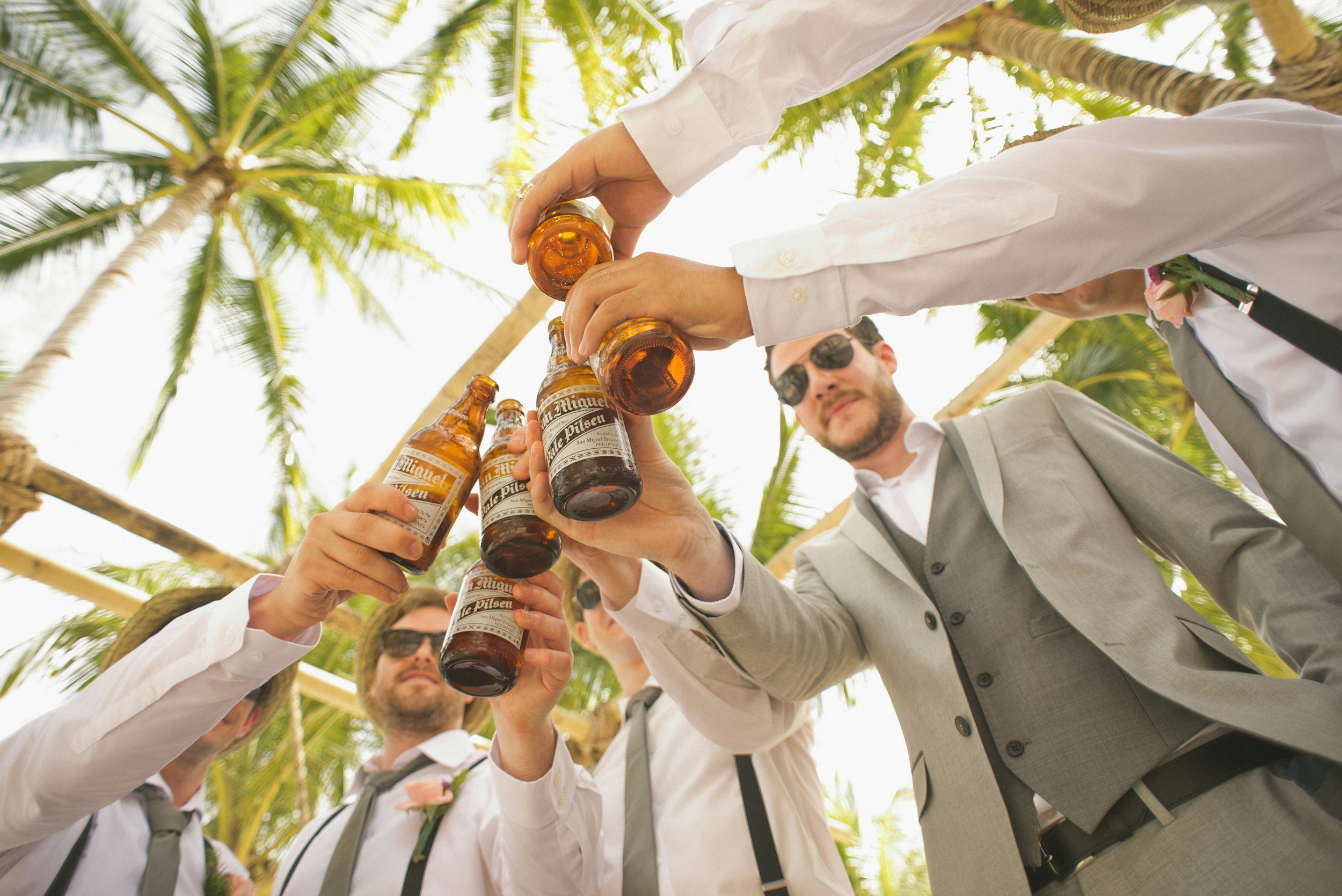 Group of men in suits toasting with beer bottles under palm trees.