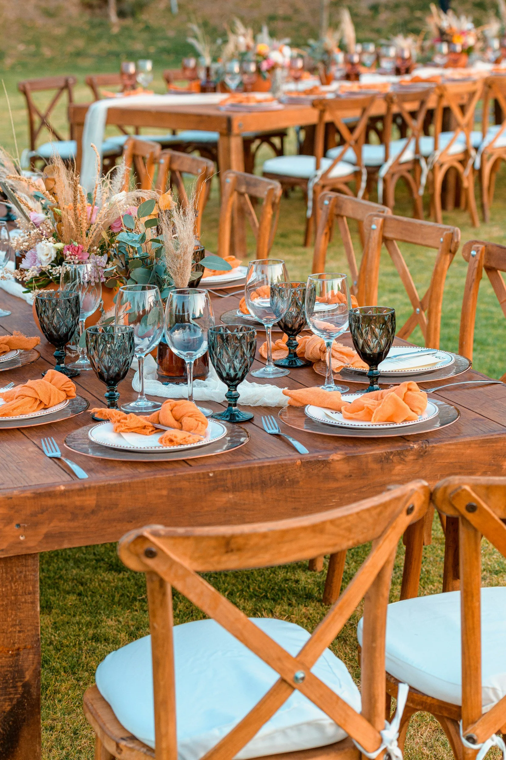 Outdoor wedding or event dining setup with wooden tables decorated with orange napkins, black and clear glassware, white plates, and floral centerpieces with dried and fresh flowers, set on green grass with matching wooden chairs.