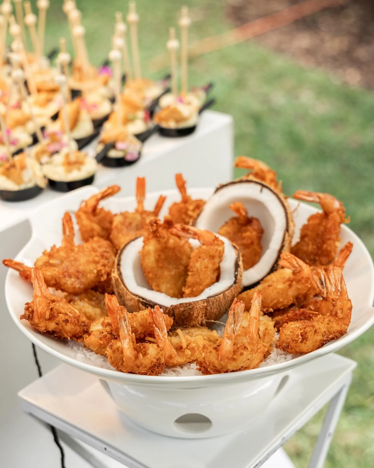 A bowl of coconut shrimp with sliced coconuts and skewered appetizers in the background.