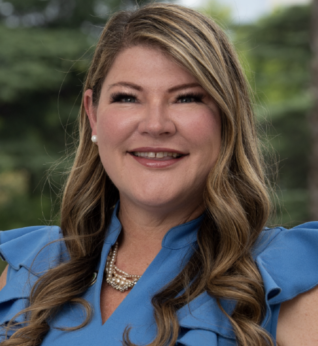 Portrait of a woman with long wavy brown hair, wearing a blue dress, pearl earrings, and a pearl necklace, smiling outdoors with trees in the background.