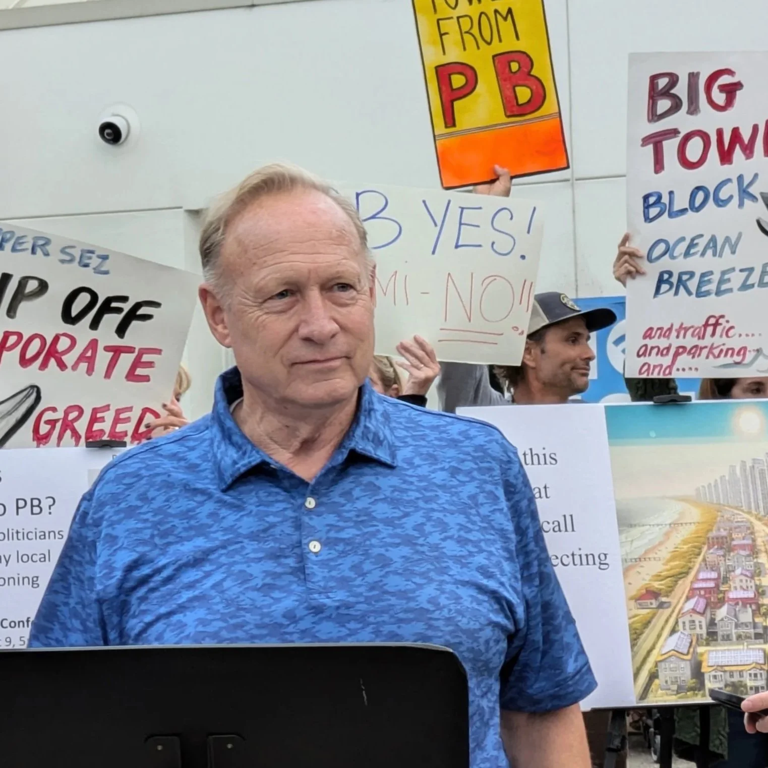 A man stands in front of a crowd holding protest signs about housing development and local control, with other protesters behind him holding colorful signs and posters.