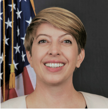 Smiling woman with short blonde hair in front of American flag.