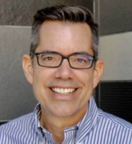 A man with short, dark hair and glasses, smiling, wearing a blue and white striped button-up shirt standing outdoors against a modern building background.