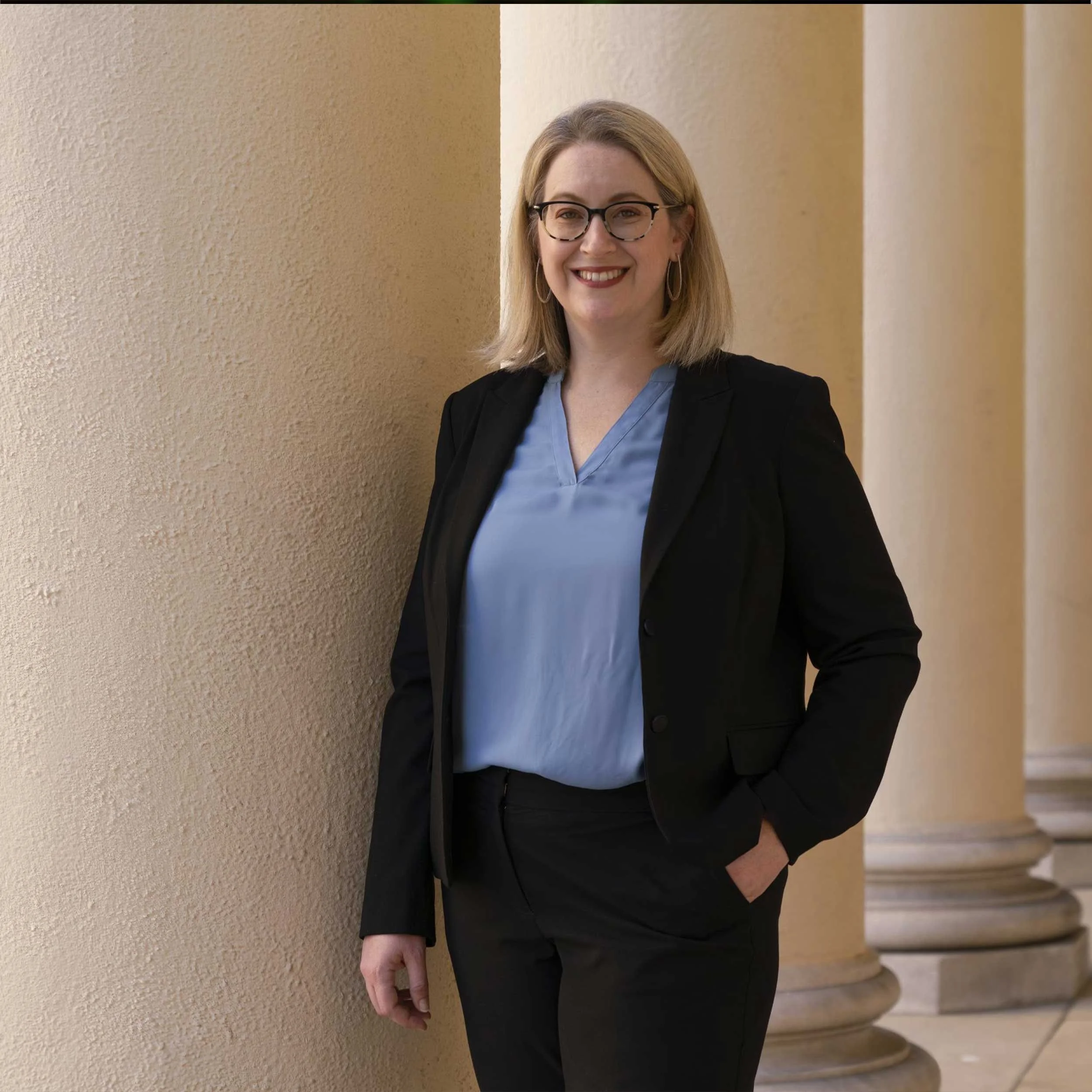 A woman with shoulder-length blonde hair, glasses, and hoop earrings, wearing a black blazer over a blue blouse, standing by a beige wall and columns, smiling at the camera.