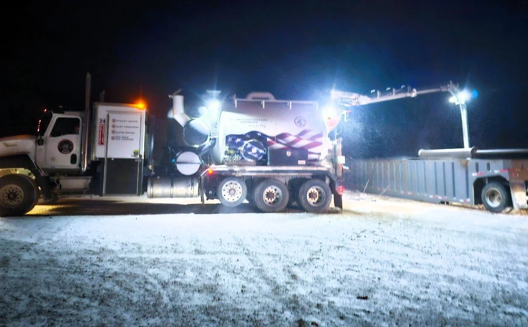 A large truck with bright lights on at night, on a snowy ground, with an extended arm or crane on the back.
