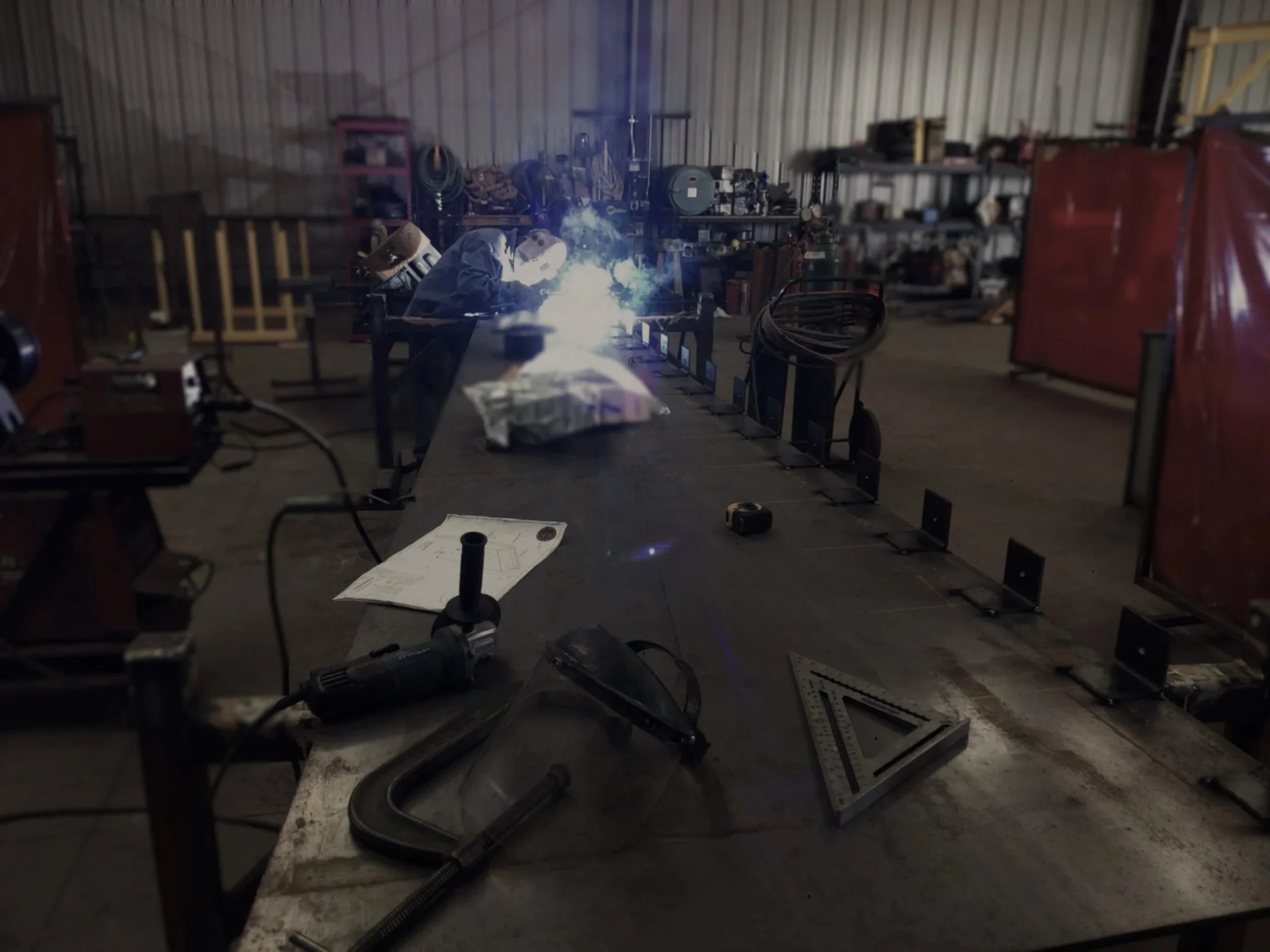 A person welding on an industrial workbench inside a workshop, with welding equipment and tools scattered around.