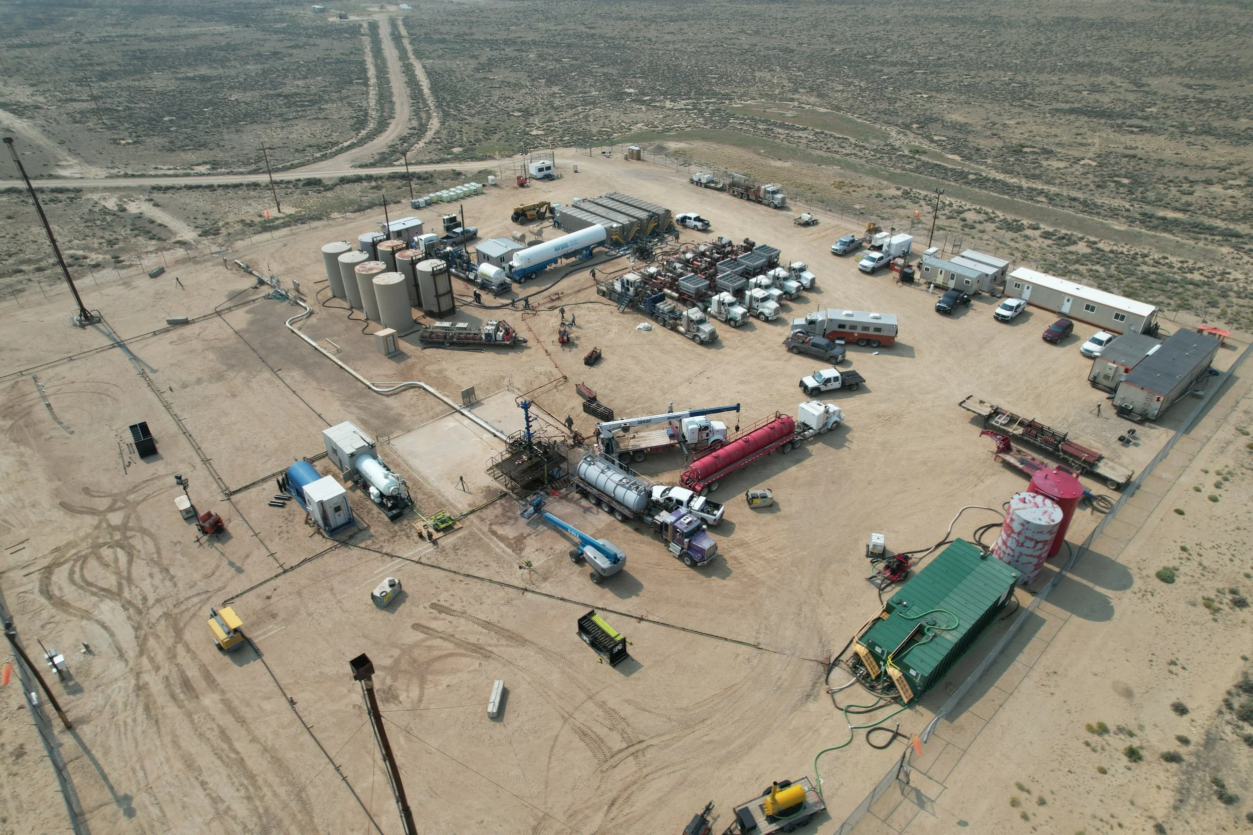 An aerial view of a temporary industrial or construction site in a desert area, with various trucks, tanks, and equipment, surrounded by dirt roads and sparse vegetation.