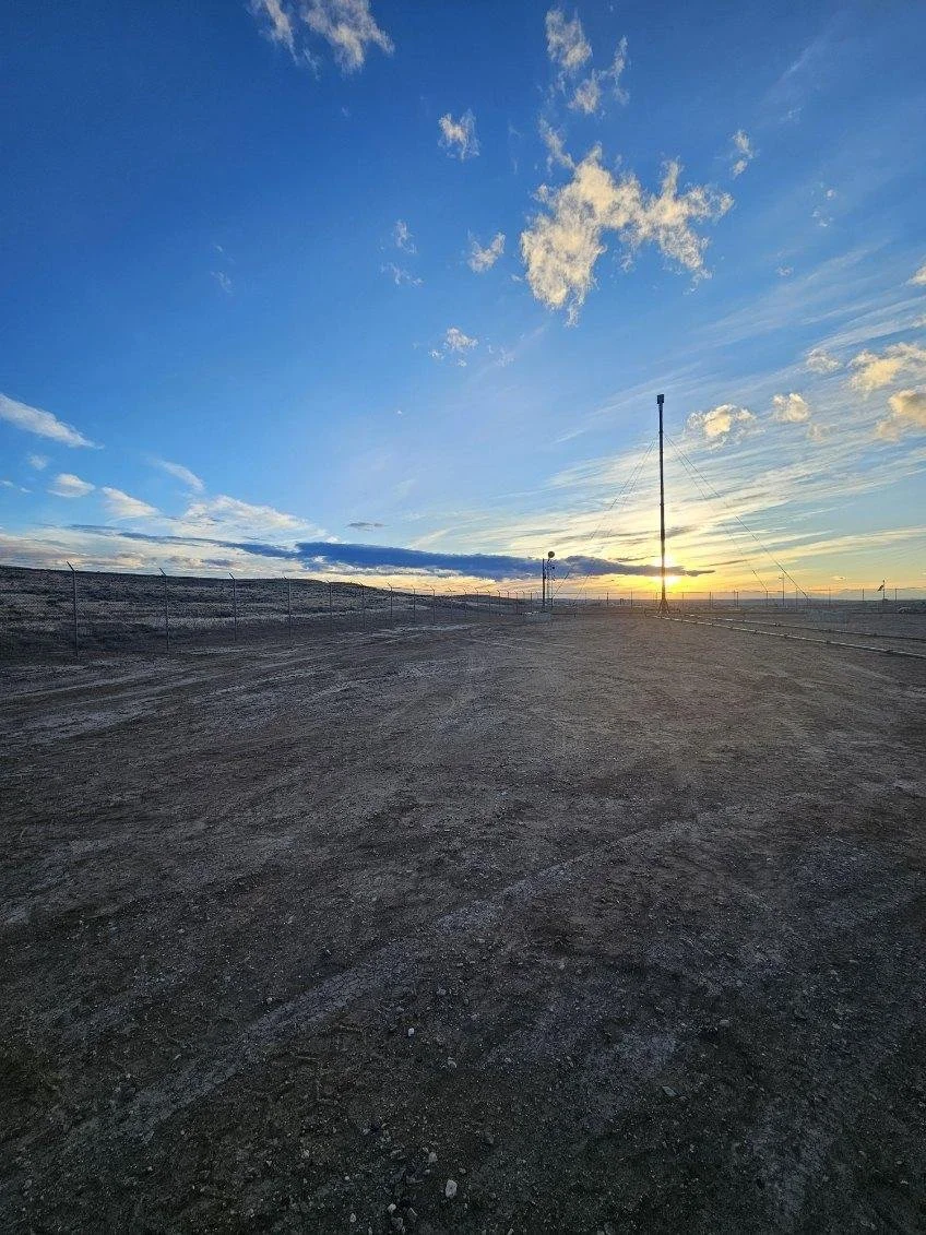 An open, dirt road with a chain-link fence to the left, power pole and lines in the distance, and the sun setting or rising on the horizon under a partly cloudy sky.
