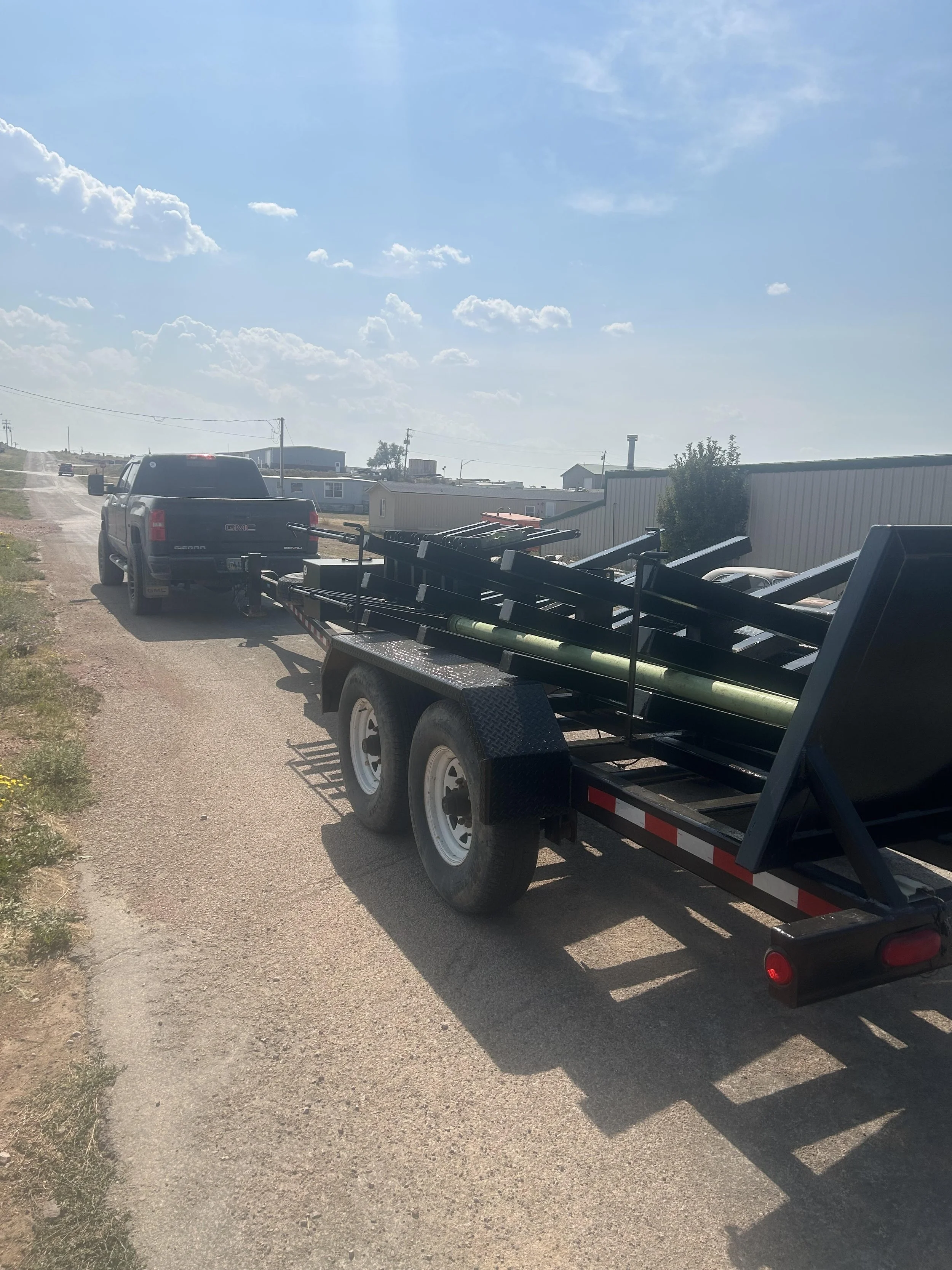 A black GMC Sierra pickup truck towing a flatbed trailer loaded with black metal framework and green pipes on a dirt road in a rural area with houses and a blue sky with clouds