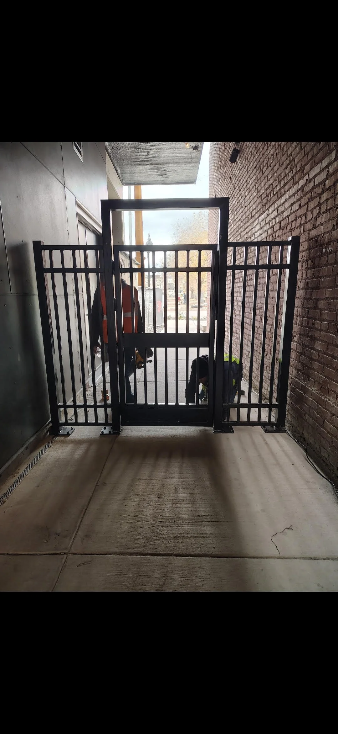 Two workers installing or repairing a black metal gate at an entrance, with one bending down and the other standing outside.