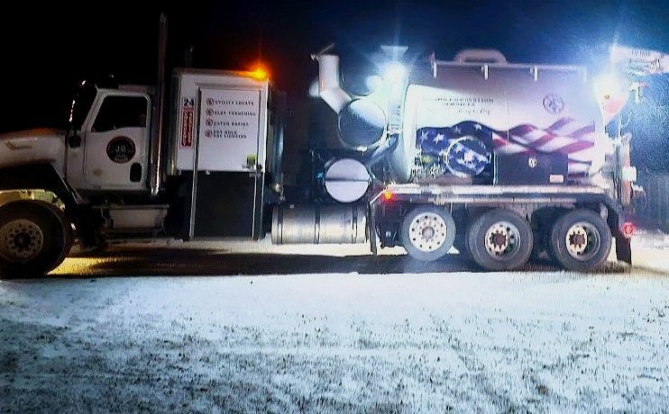 A large truck with a commemorative American flag and motorcycle painted on the side, parked on a snow-covered road at night, with headlights and an orange warning light on.