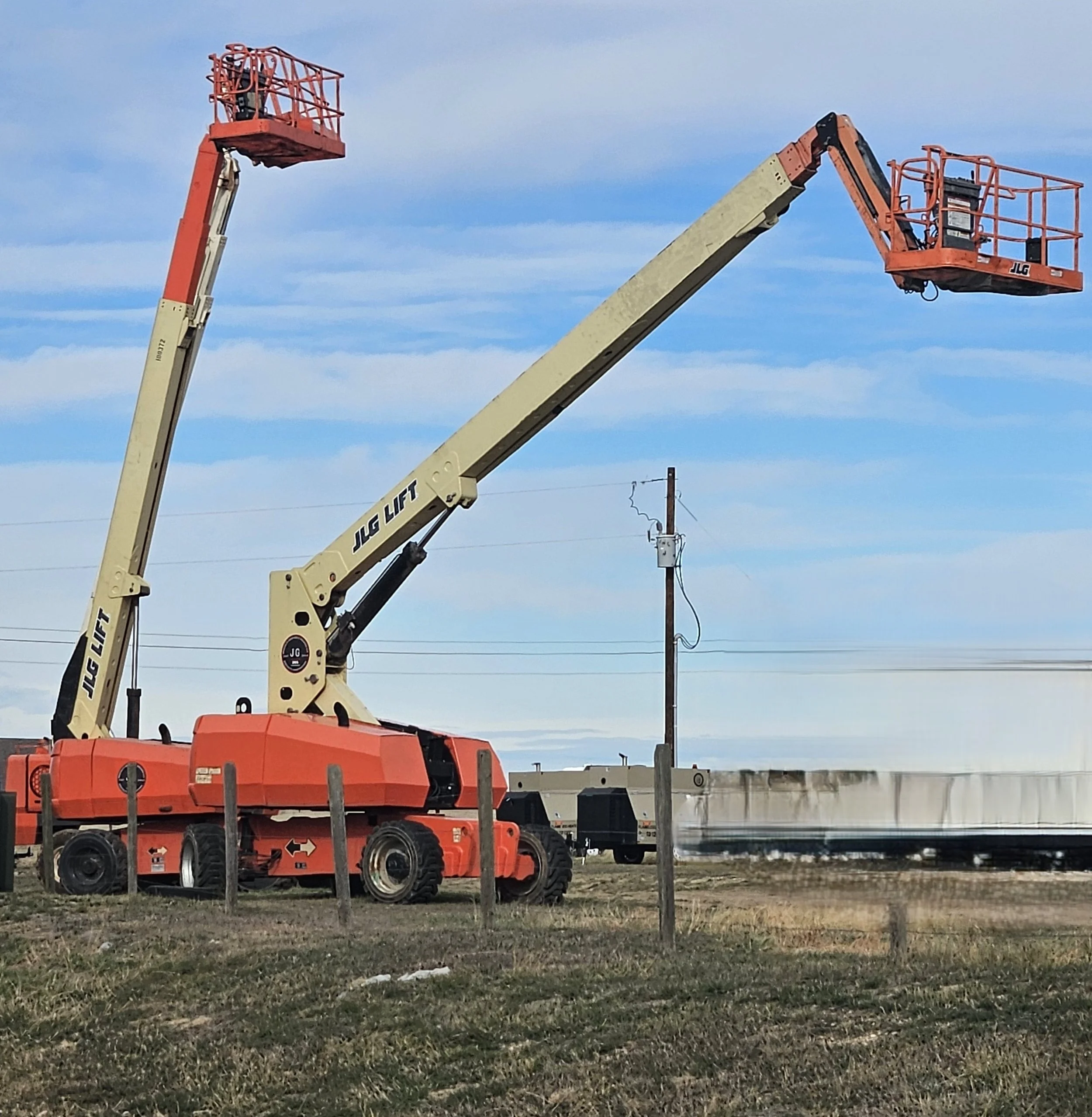 A large orange and beige JLG lift with extended arms and two aerial work baskets, positioned on a grassy area near a train track and utility poles under a blue sky.