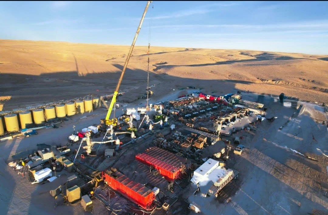 A large industrial site in a desert landscape with yellow storage tanks, various equipment, and multiple vehicles, including trucks and trailers, under a clear blue sky.