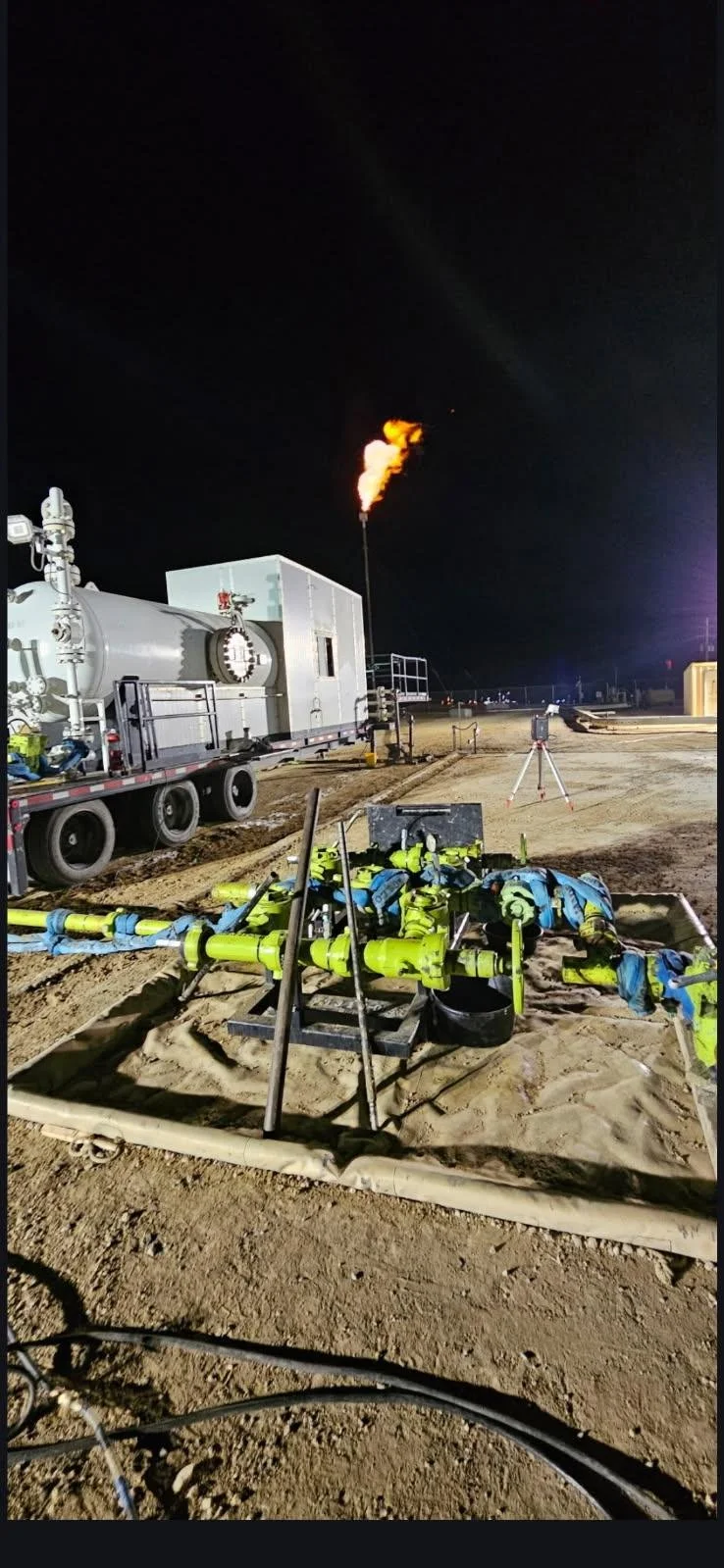 Nighttime scene of industrial equipment and pipes on a construction or industrial site, with a flare stack emitting fire into the dark sky. flare stack and 1440psi high stage