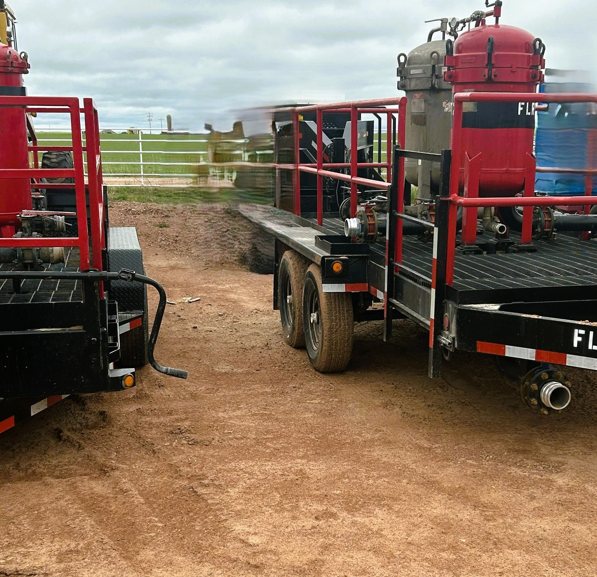 Two fire trucks with large red water tanks parked on a dirt ground, with a green field and cloudy sky in the background.