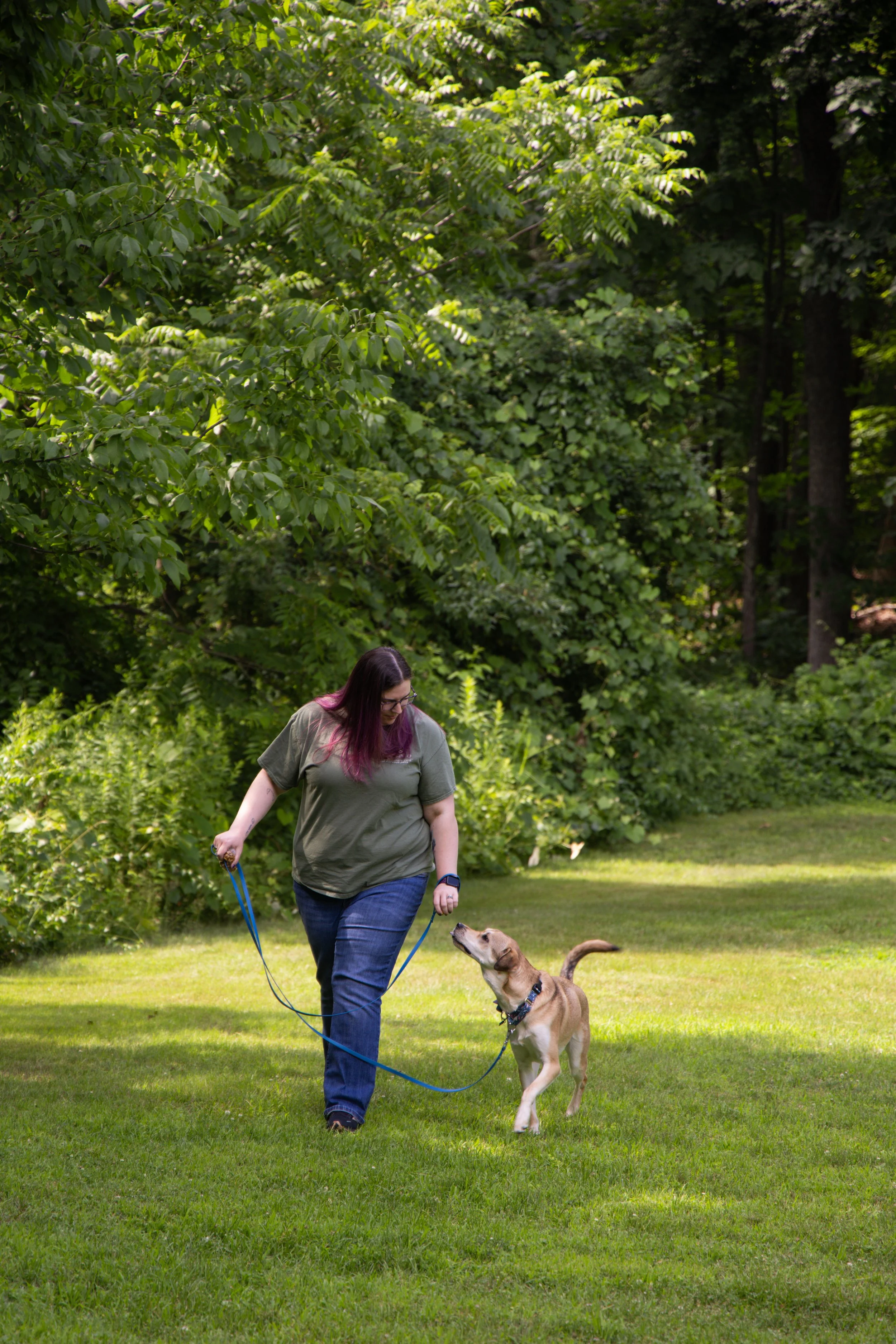 A woman with purple hair and glasses walking a dog on a grassy field in a wooded area.