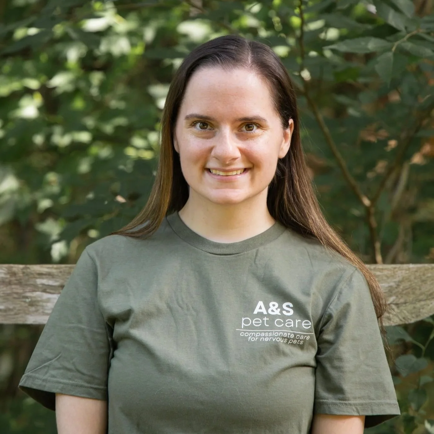 A woman kneeling on grass beside a brown dog on a leash, outdoors in a park with trees in the background, smiling at the camera.