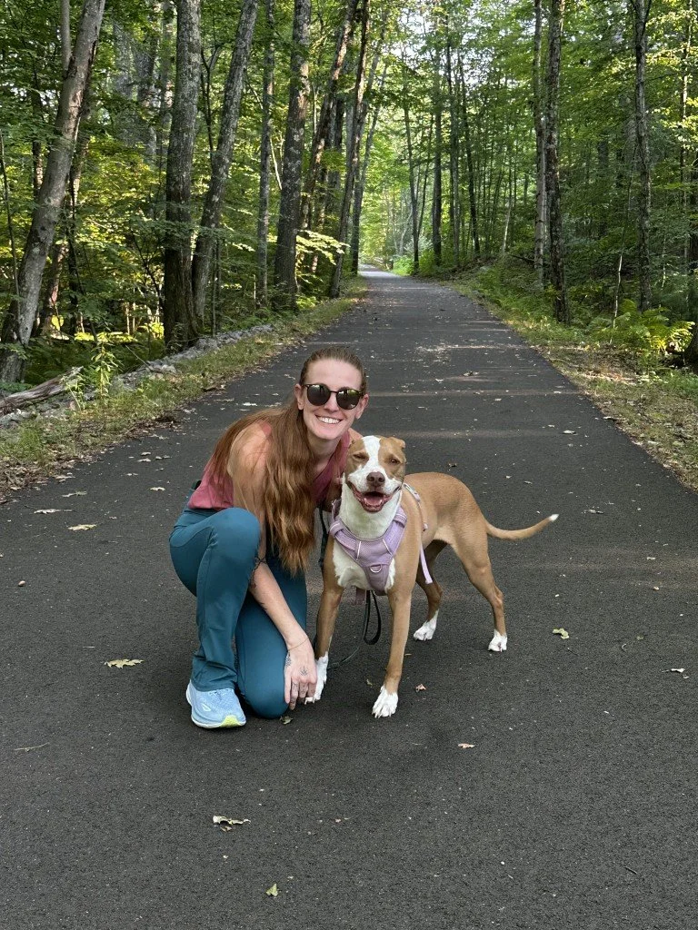 Woman with brown hair wearing sunglasses, a purple knit hat, and a gray plaid fleece jacket, smiling while holding a white cat on her shoulder during winter on a rural road.