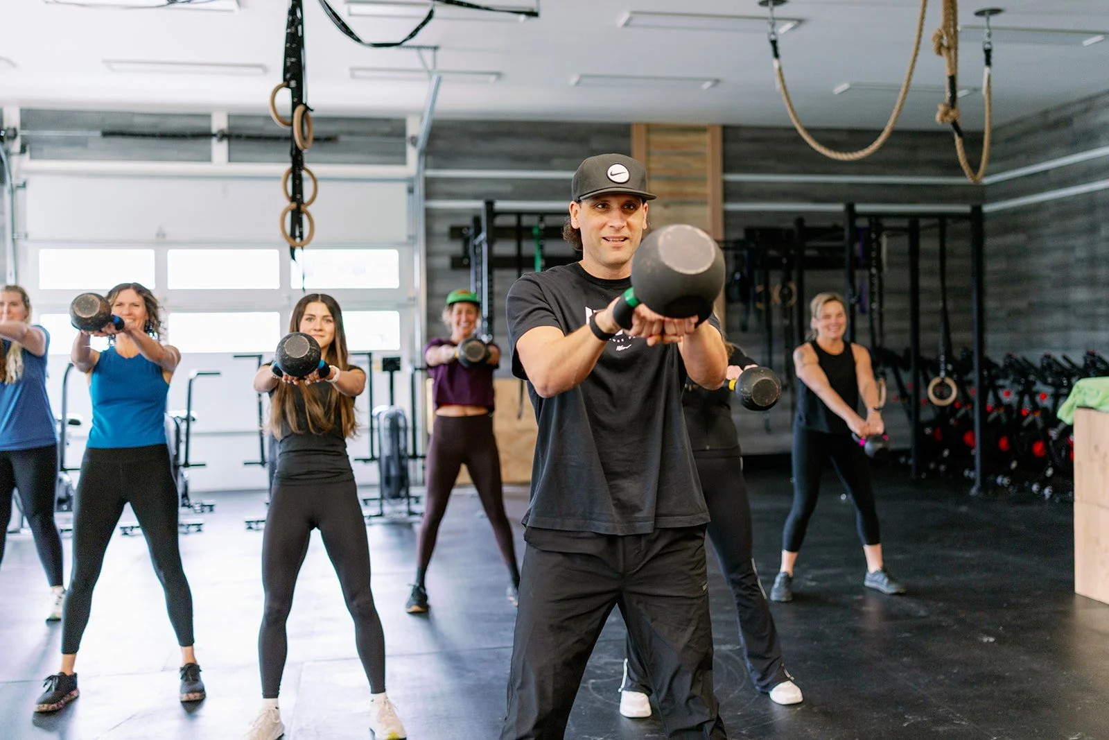 A man in a black tank top and shorts pulls on a thick battle rope at a gym, with weightlifting equipment and a woman with a child in the background.