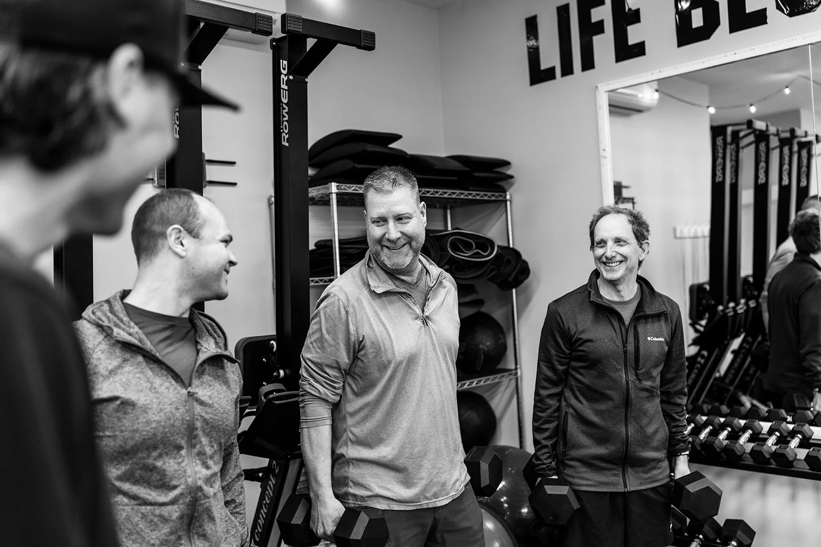 A man in a black tank top and shorts pulls on a thick battle rope at a gym, with weightlifting equipment and a woman with a child in the background.