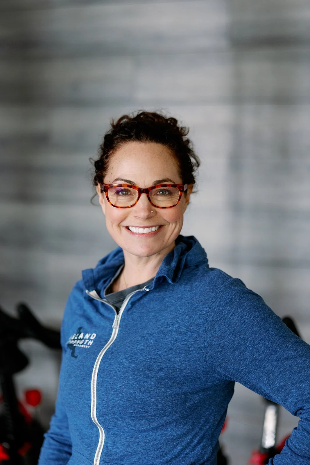 A smiling woman in a blue t-shirt with the words 'Island Strength + Movement' and a silhouette of California, standing in a gym.