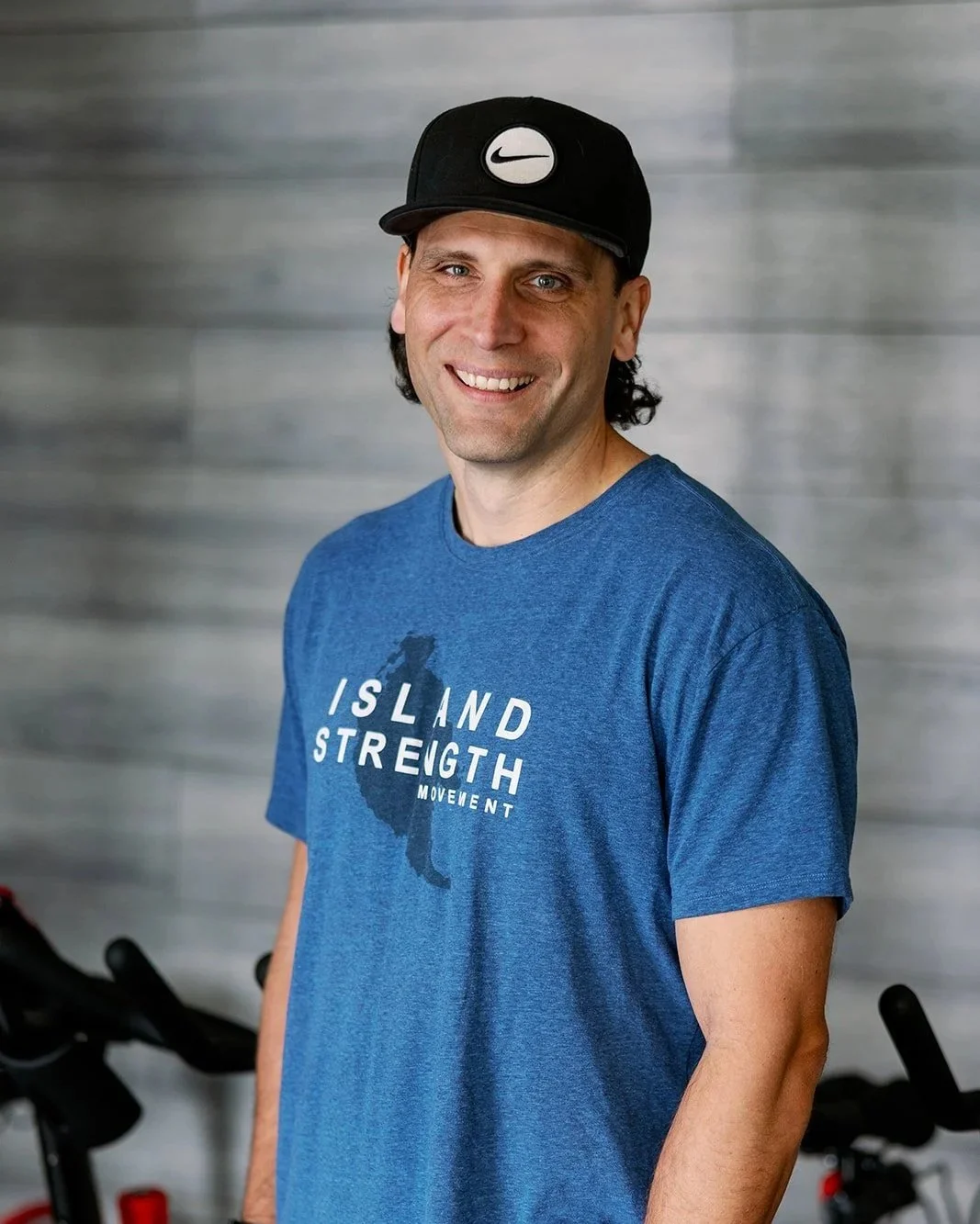 A smiling man in a blue t-shirt with the text 'ISLAND STRENGTH MOVEMENT' and a graphic of an island, standing in a gym.
